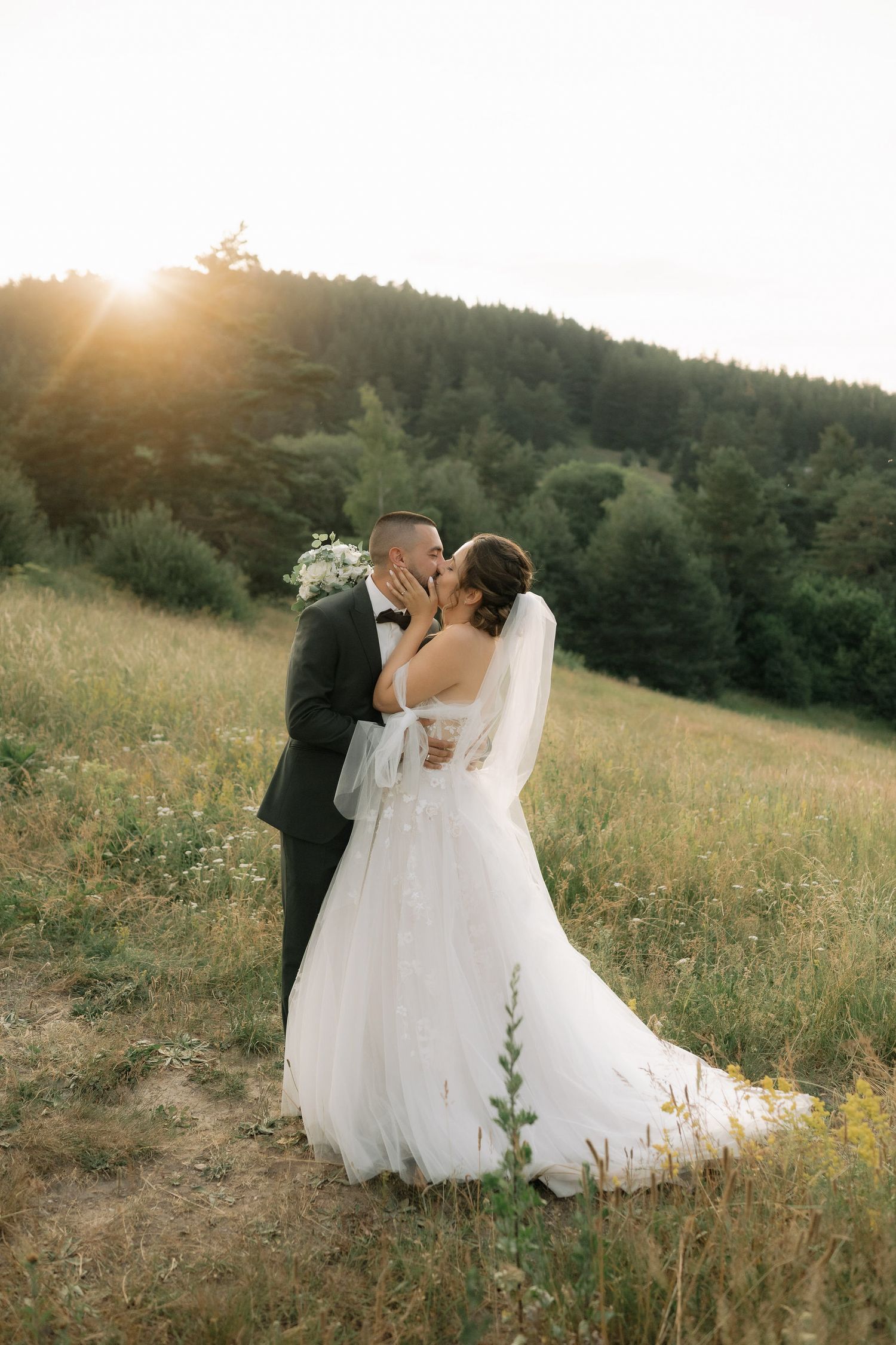 Newlyweds share an intimate moment in a golden field at sunset with evergreen trees in the distance.