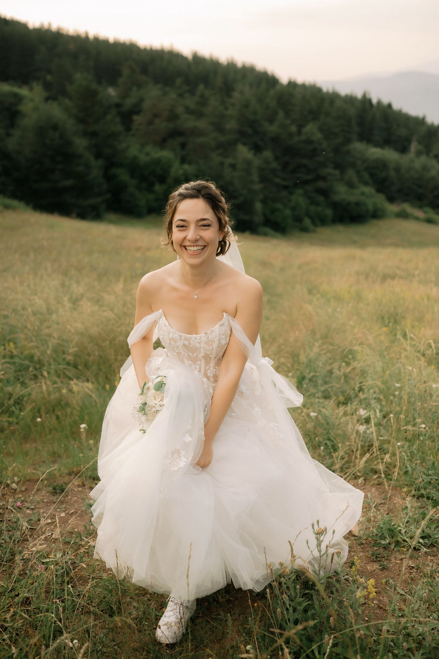 A bride in a flowing white wedding dress sits gracefully in a meadow with evergreen trees in the background.