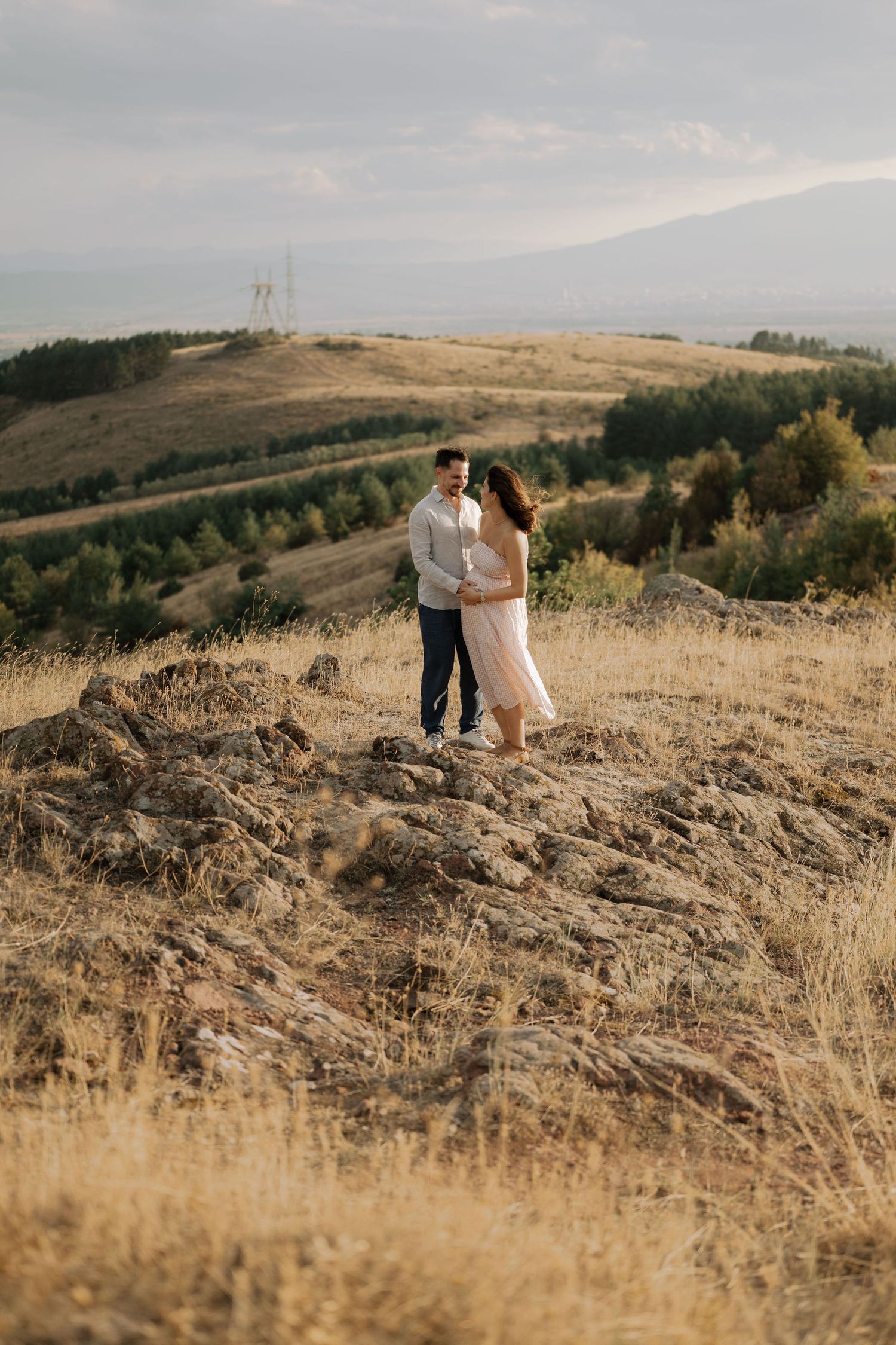 A couple embraces on a golden hillside overlooking a scenic mountain landscape during sunset.
