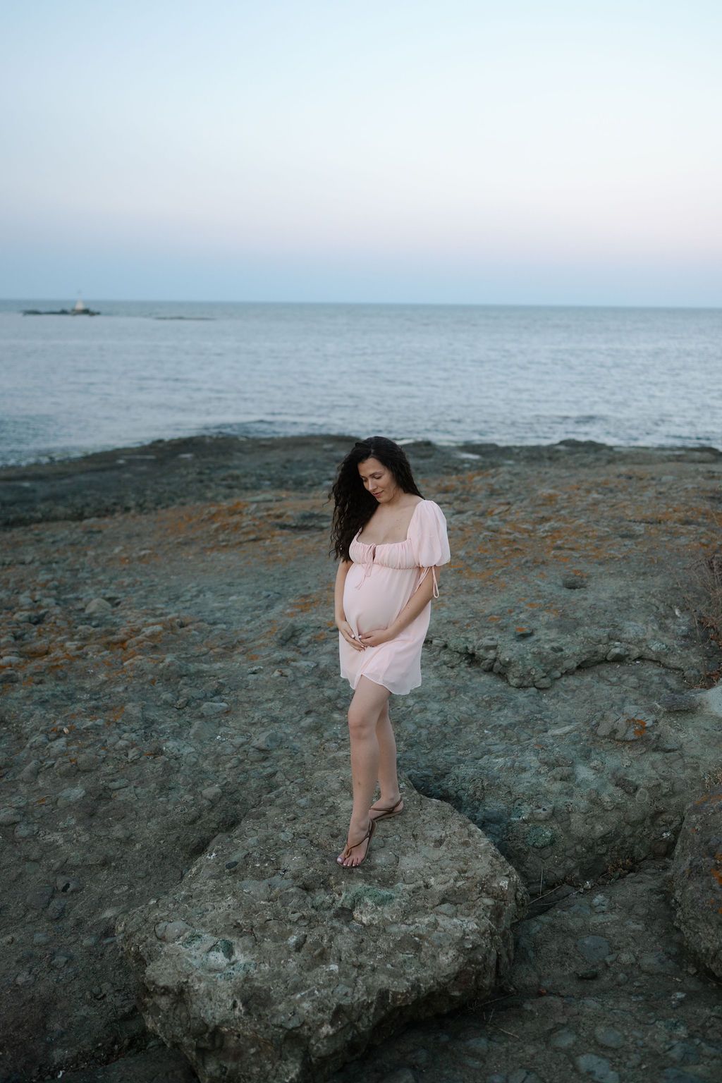 A pregnant woman in a pink dress stands on coastal rocks at sunset overlooking a calm ocean.