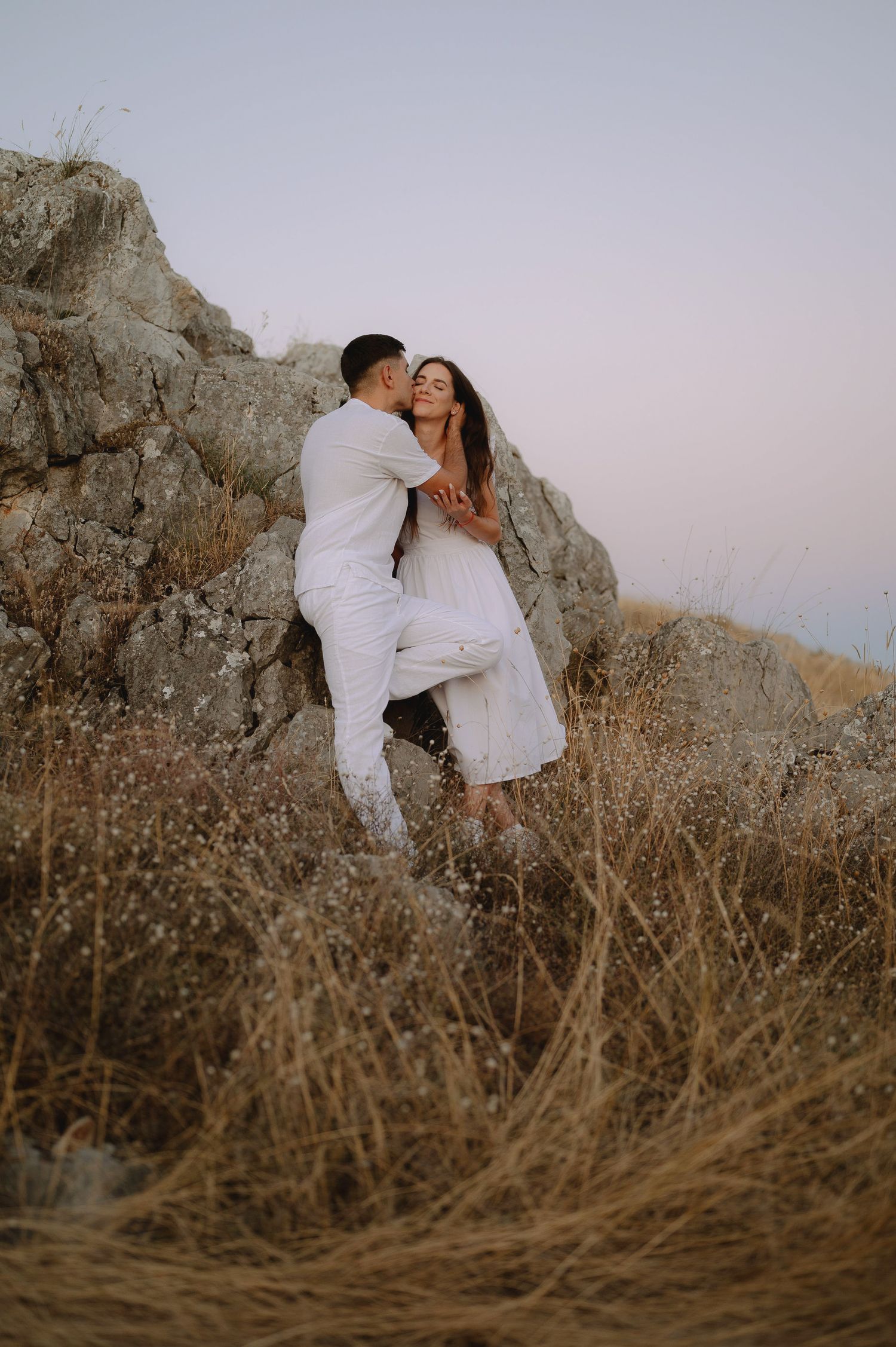 A couple in white outfits embrace on a rocky hillside during sunset among dry grass and boulders.