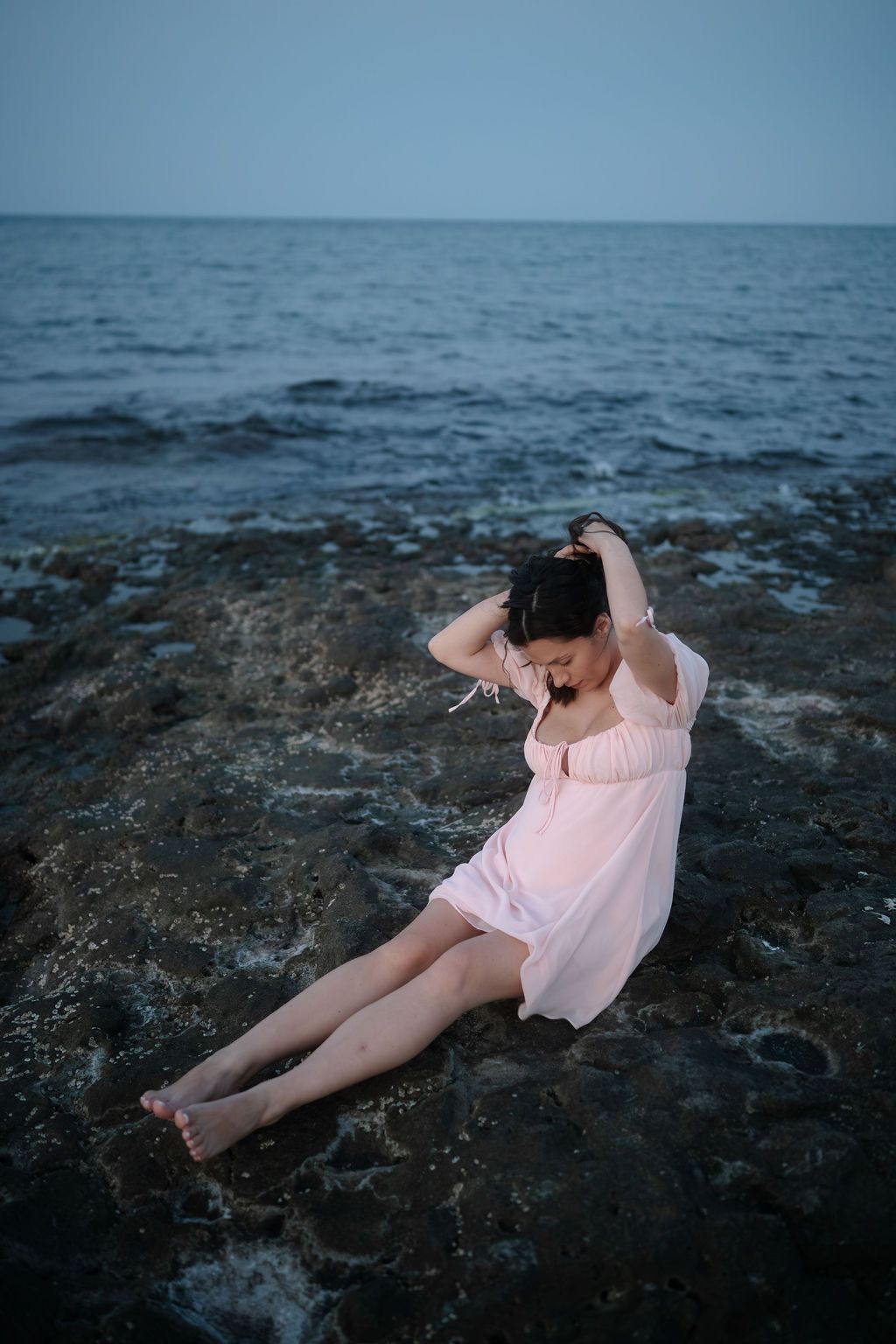 A person in a pink dress reclines on dark coastal rocks at dusk with the ocean in the background.