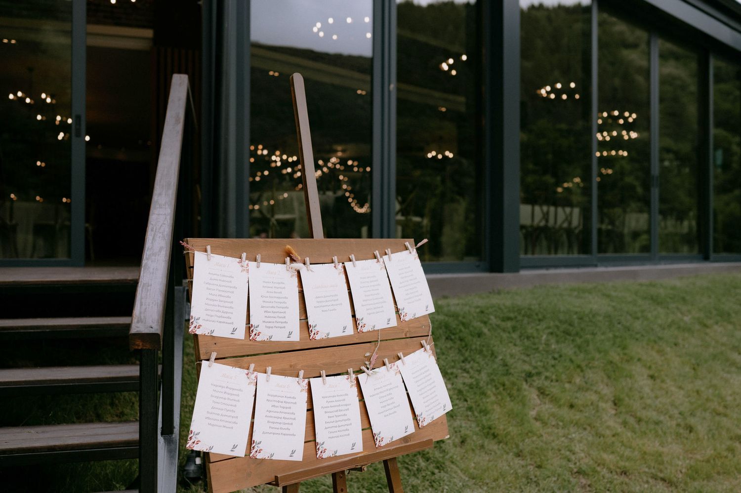 A rustic wooden seating chart with paper cards hanging from twine displays wedding guest table assignments at an outdoor venue.