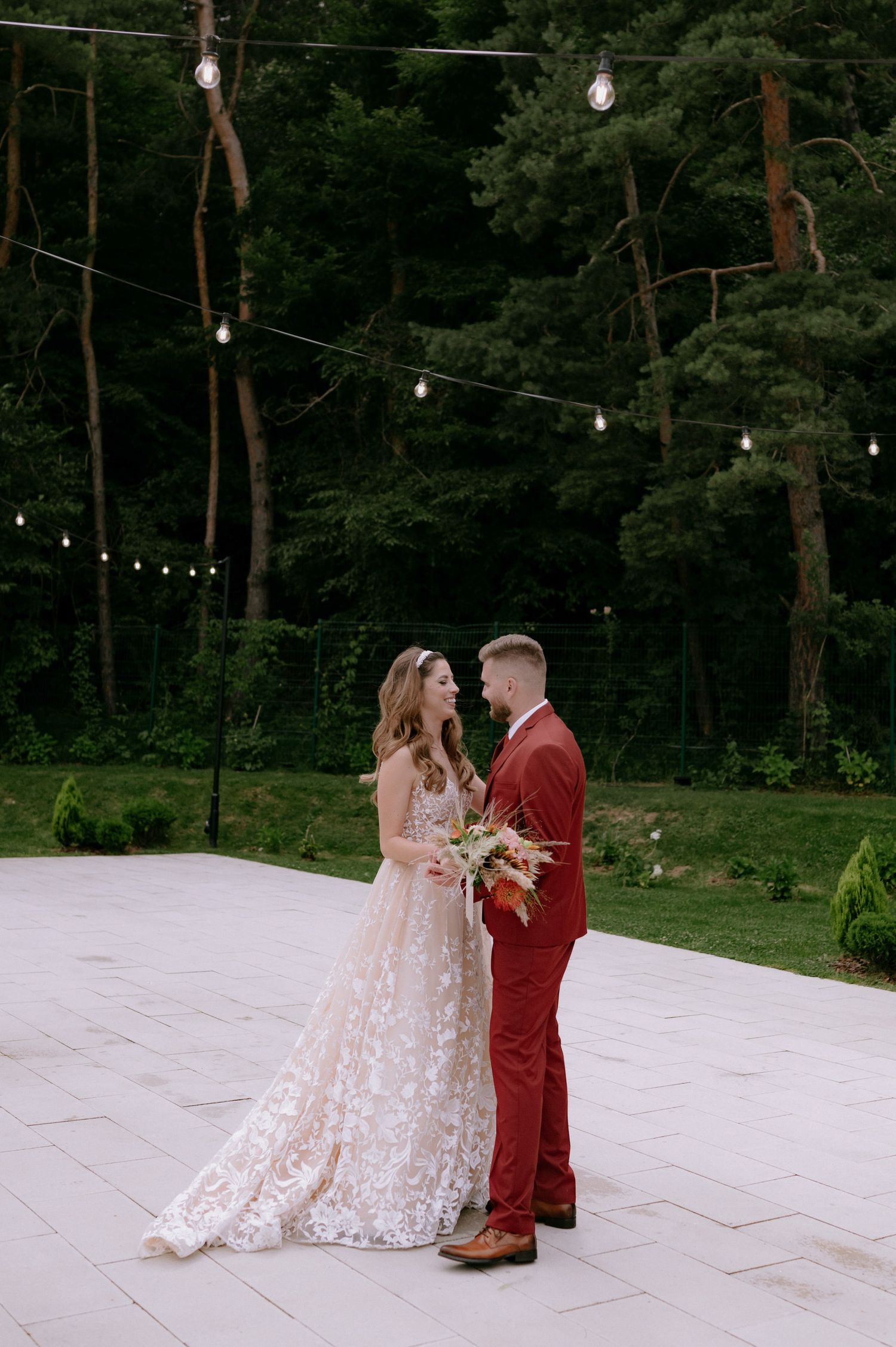 A couple shares an intimate moment on a white stone patio surrounded by string lights and tall pine trees in the background.