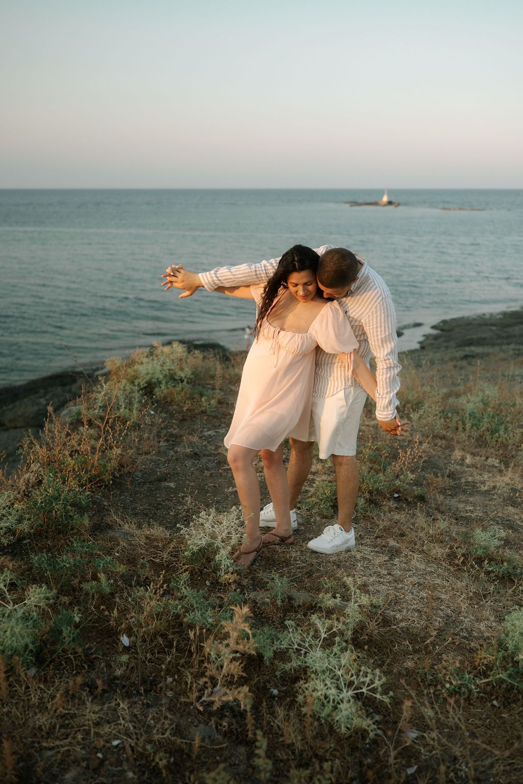 A couple in white outfits shares an intimate moment on a coastal cliff overlooking the ocean at sunset.