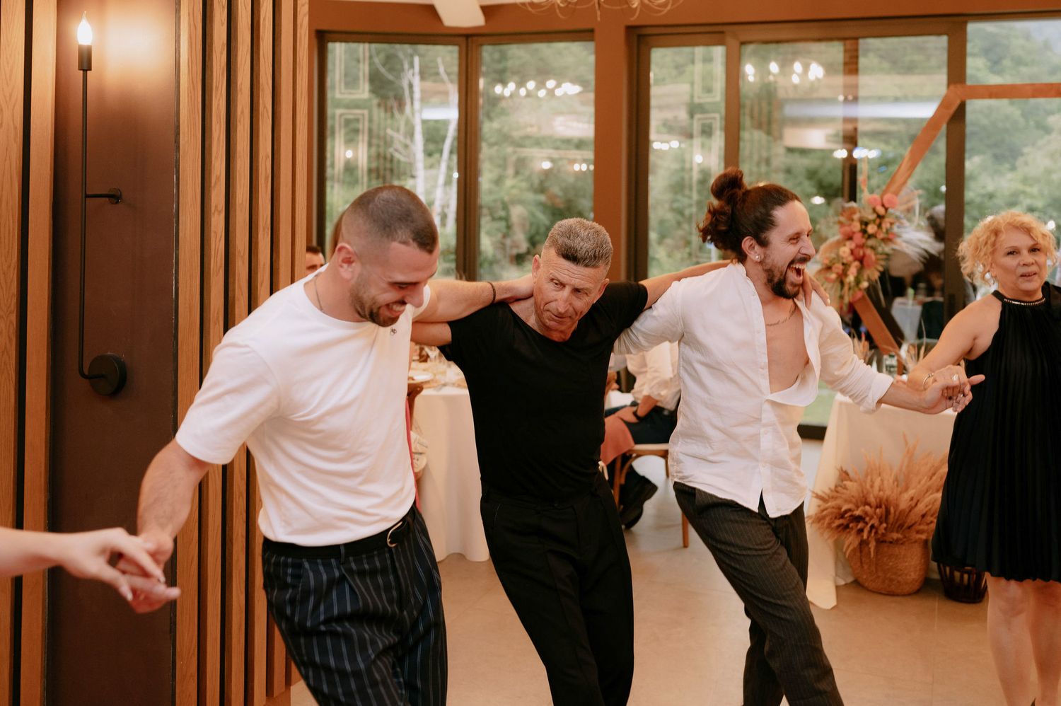 People dancing and celebrating at an indoor event with warm lighting and glass windows in the background.