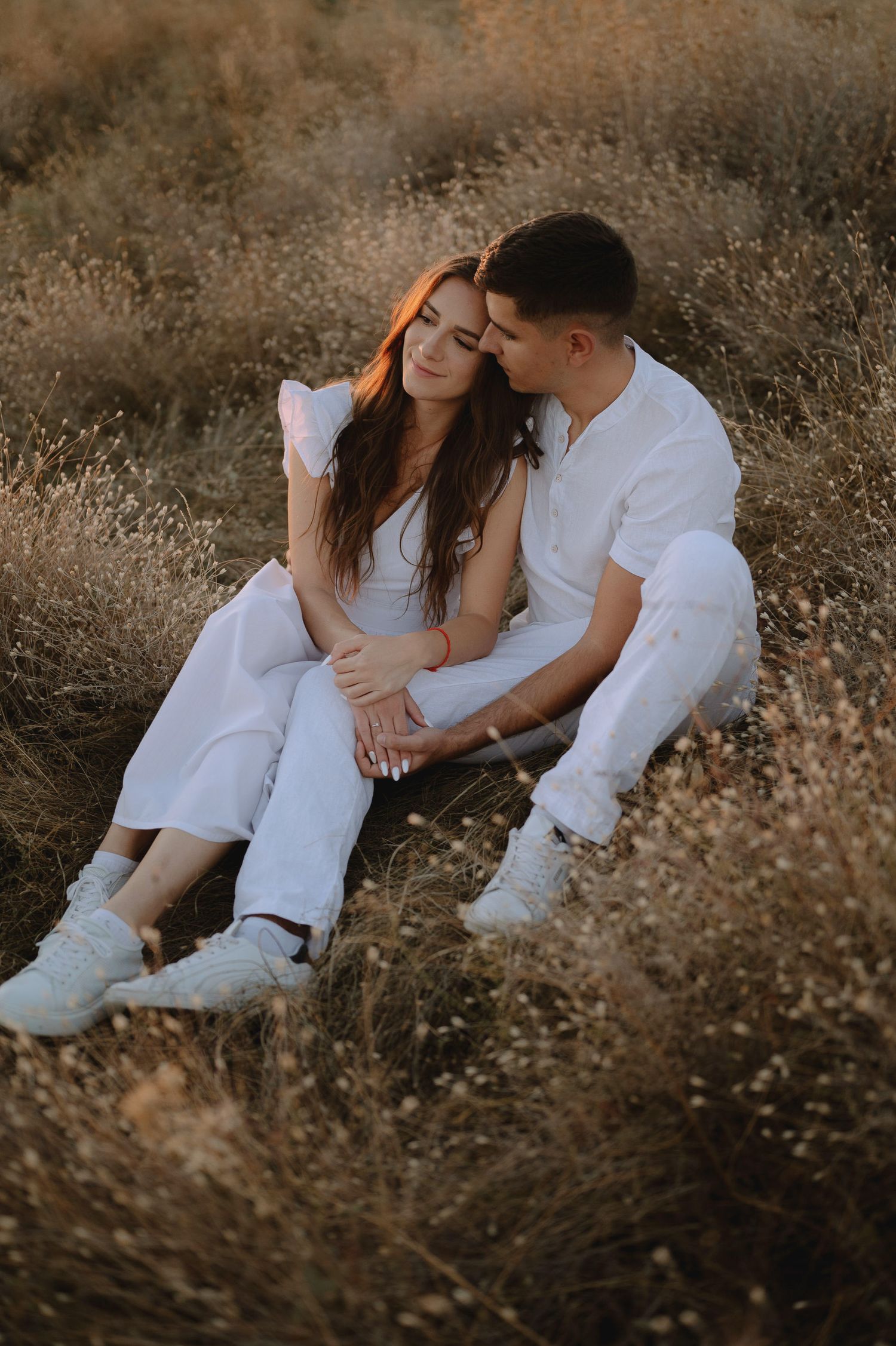 A couple in white clothing sits intimately together in a field of dried grass during golden hour.