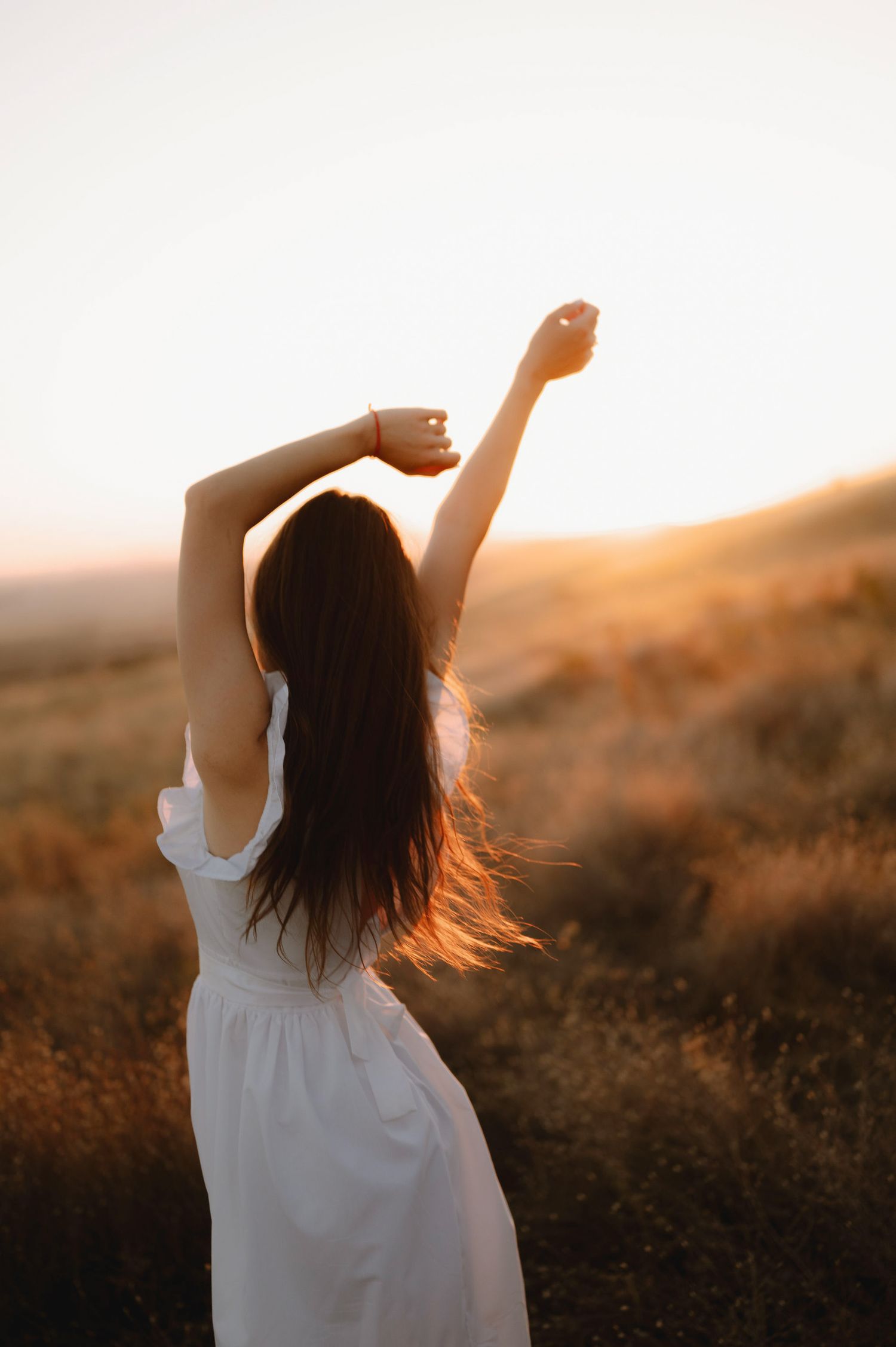 Silhouette of a person in a white dress stretching arms up during golden sunset in a grassy field.