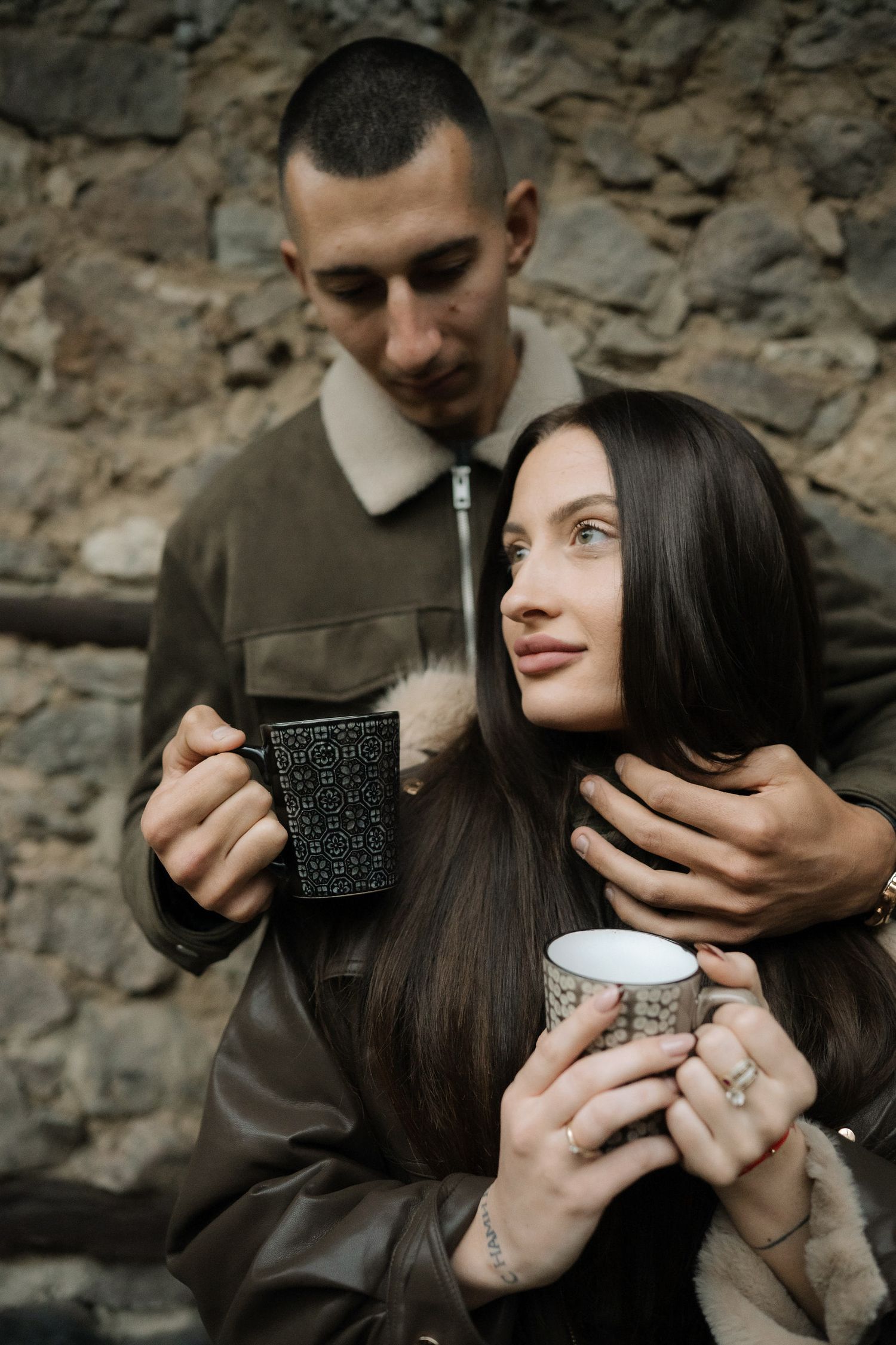 A couple shares an intimate moment while holding coffee mugs against a rustic stone wall background.