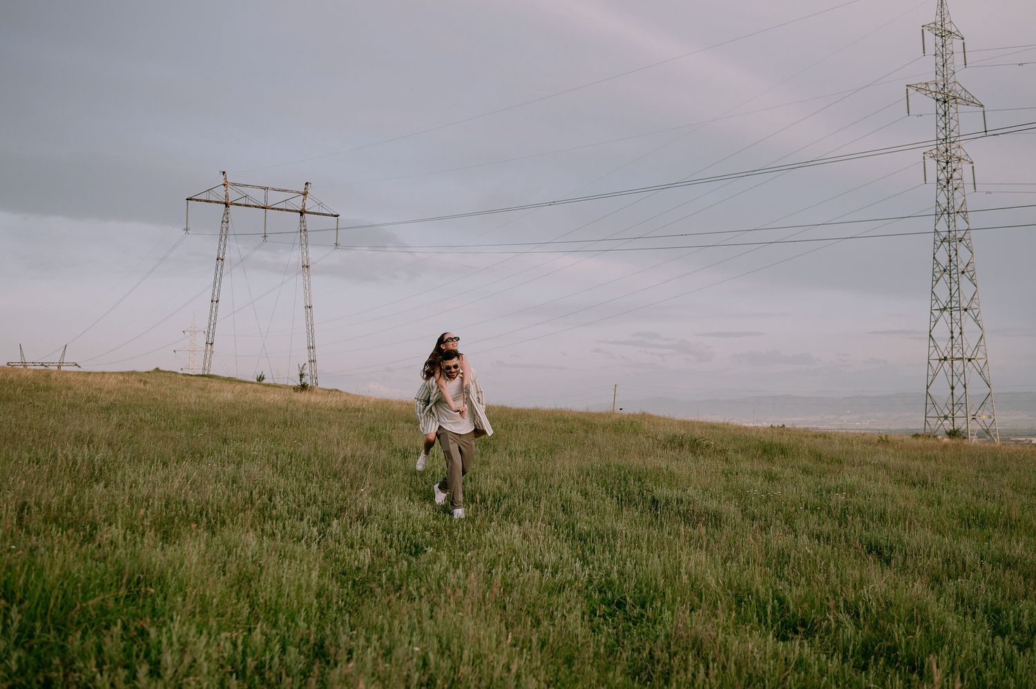 Person walking through a green meadow with power transmission towers and lines against a cloudy sky.