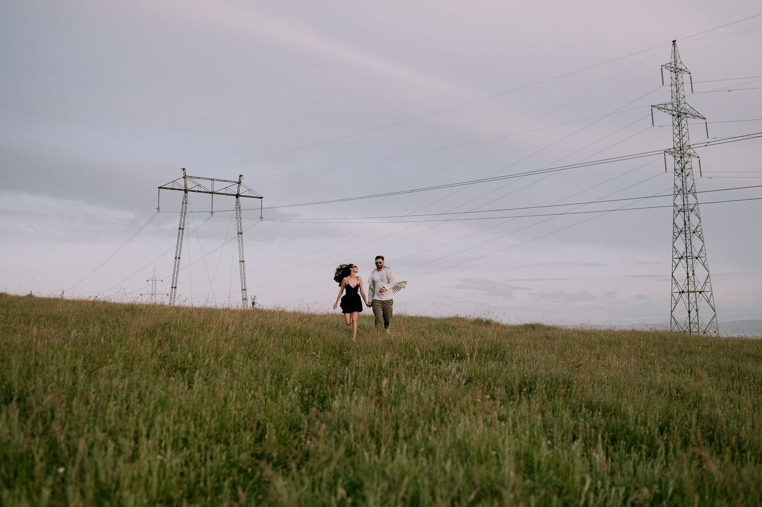 Two figures walk along a grassy hilltop beneath power transmission lines on an overcast day.