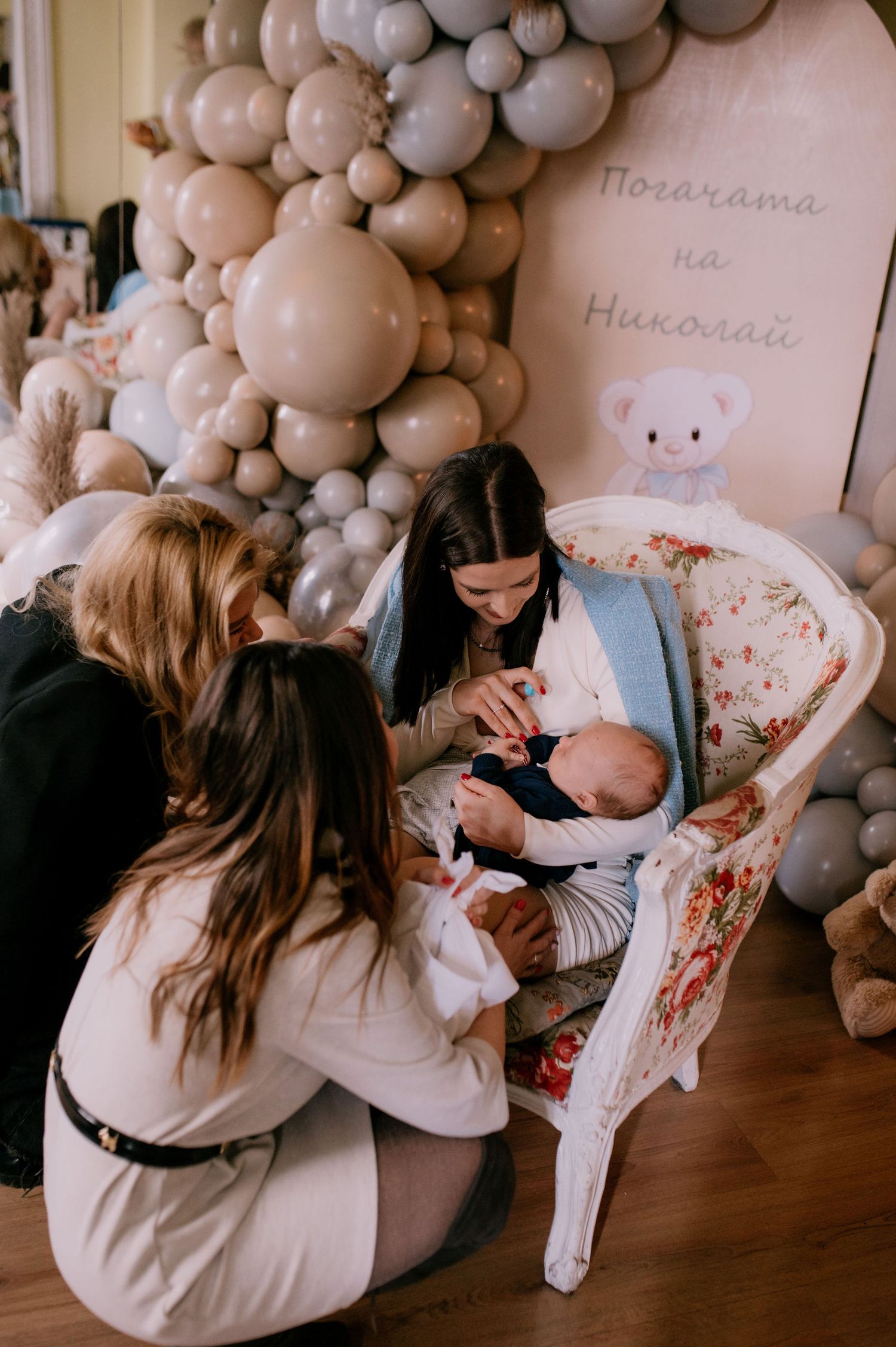 A cozy scene with a baby in a chair surrounded by gray balloon decorations and people gathered around.