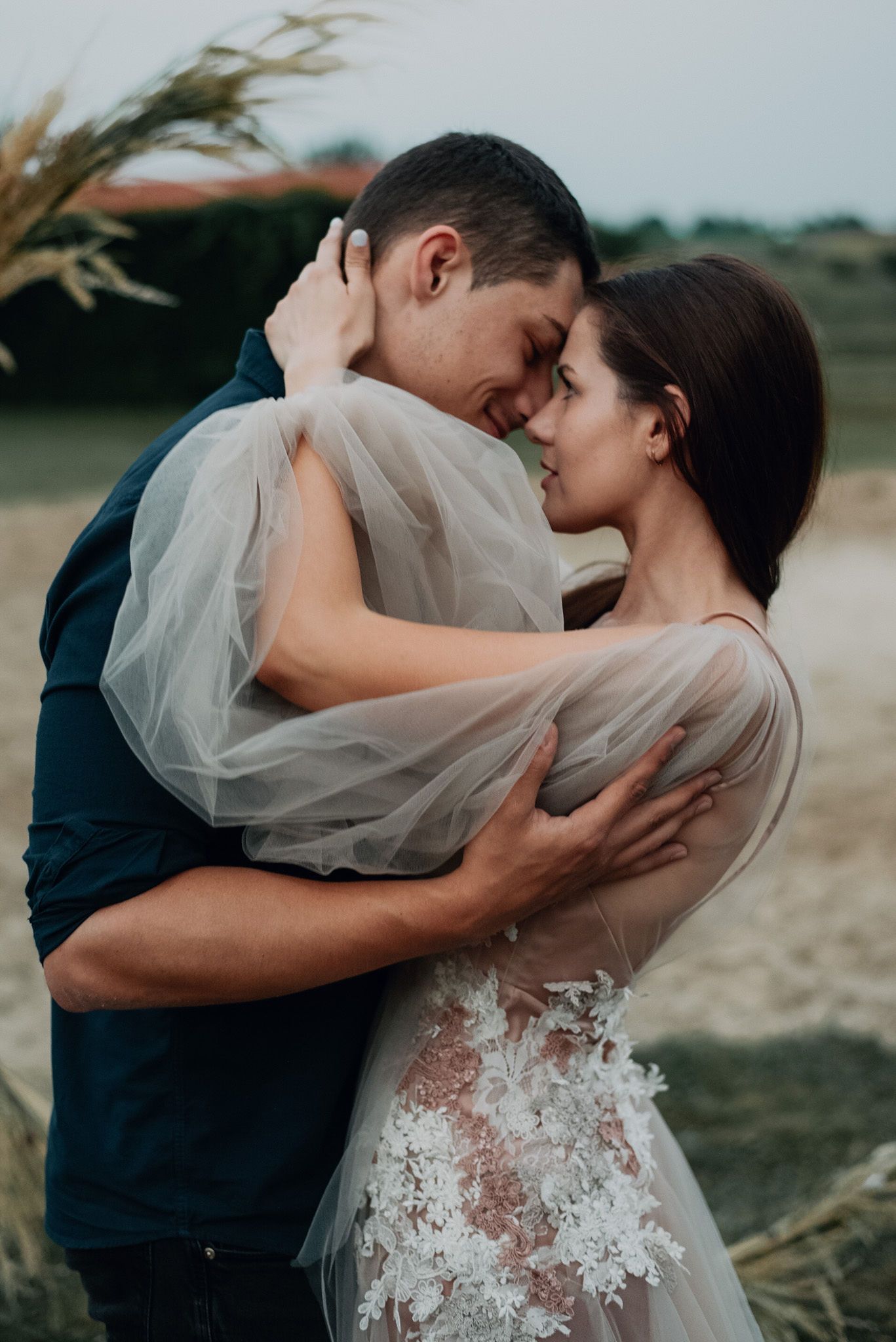 A romantic wedding couple shares an intimate kiss while the bride's veil flows in the wind at an outdoor ceremony.