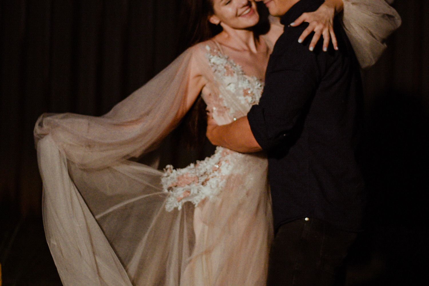 A couple dances intimately in a moody setting, with the bride wearing a flowing sheer dress with floral appliques.