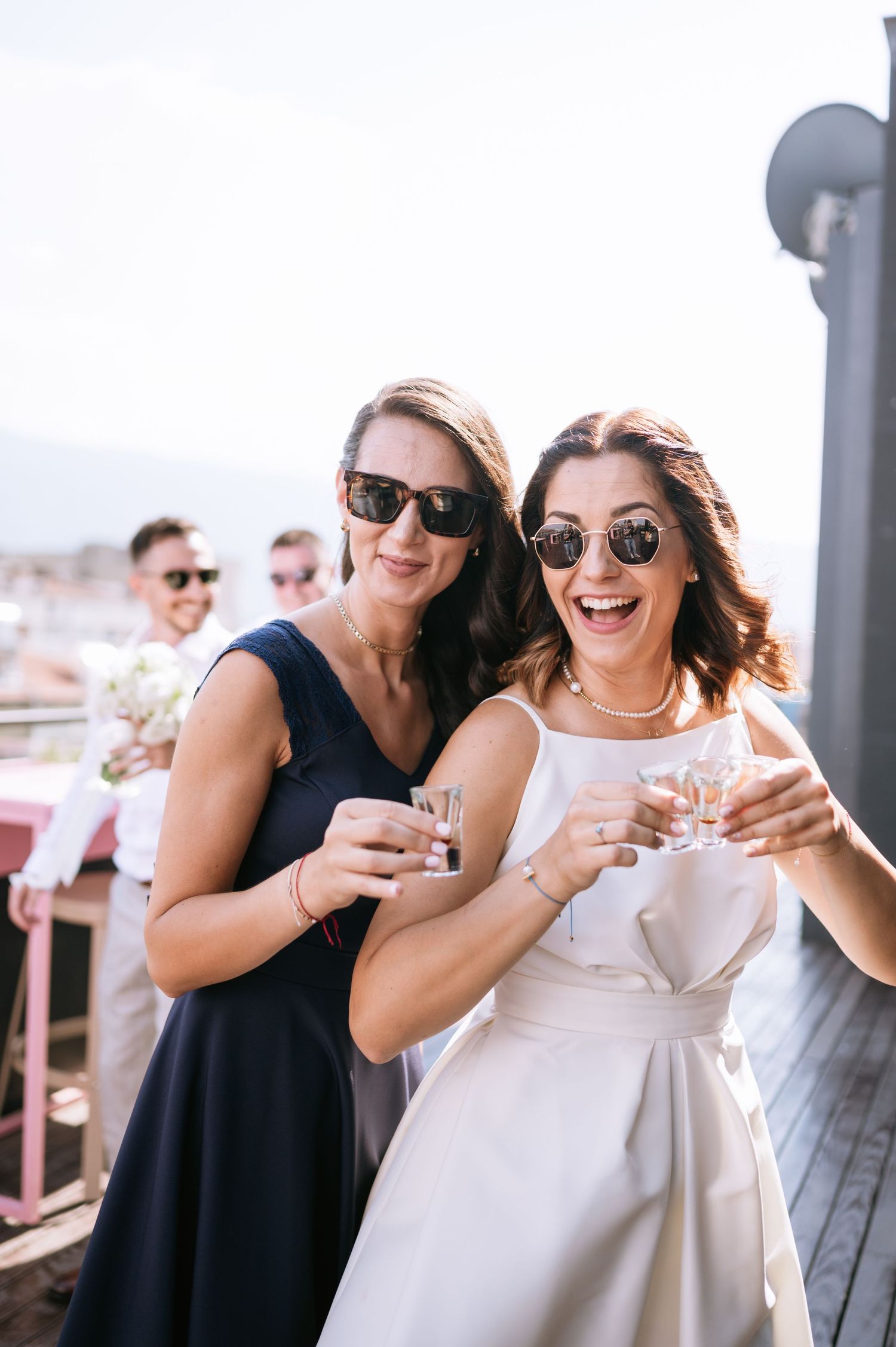 Friends laughing and celebrating together at an outdoor rooftop party while wearing sunglasses.
