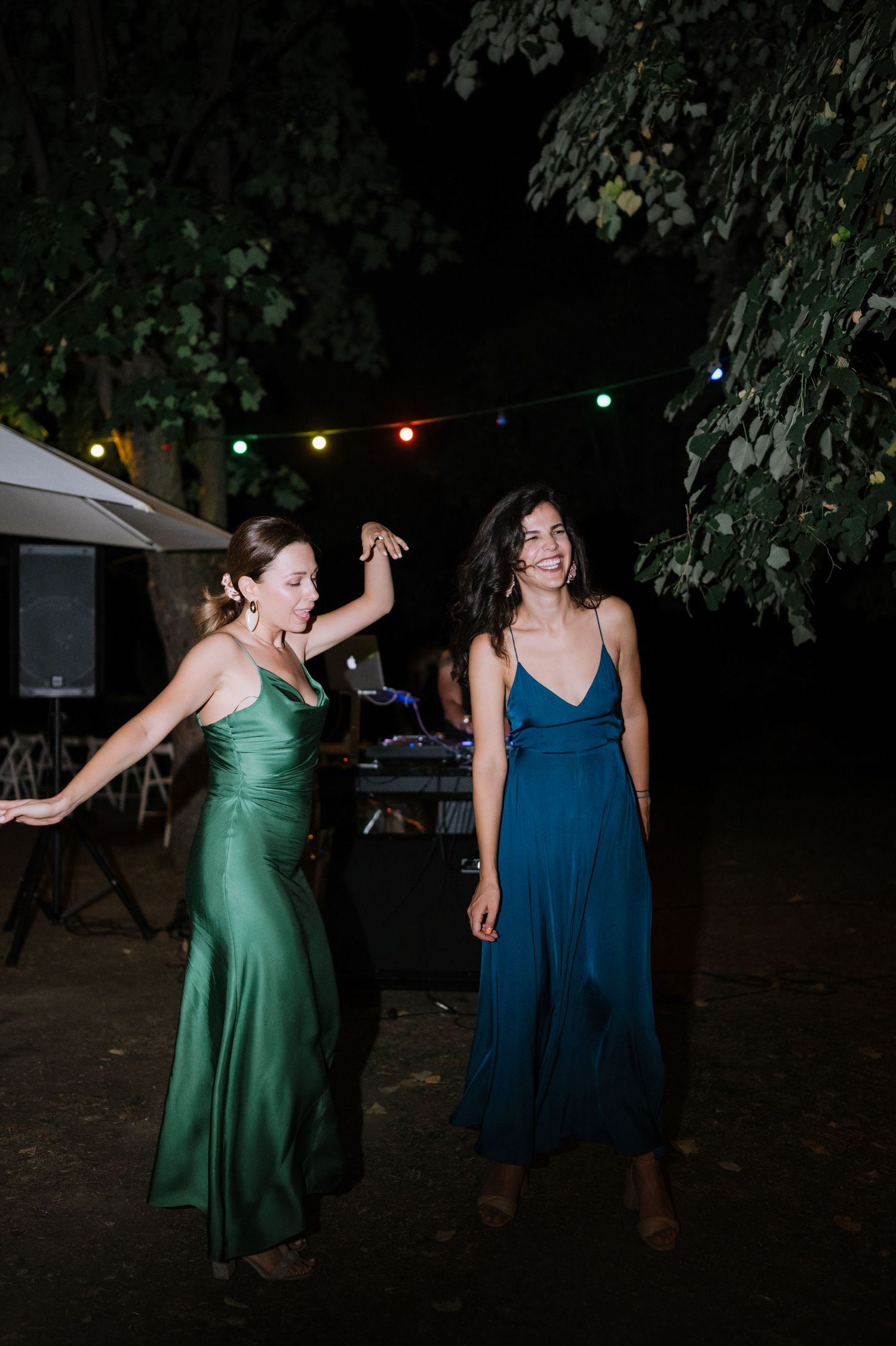 Two women in green and navy dresses dance together at a nighttime outdoor party with string lights overhead.