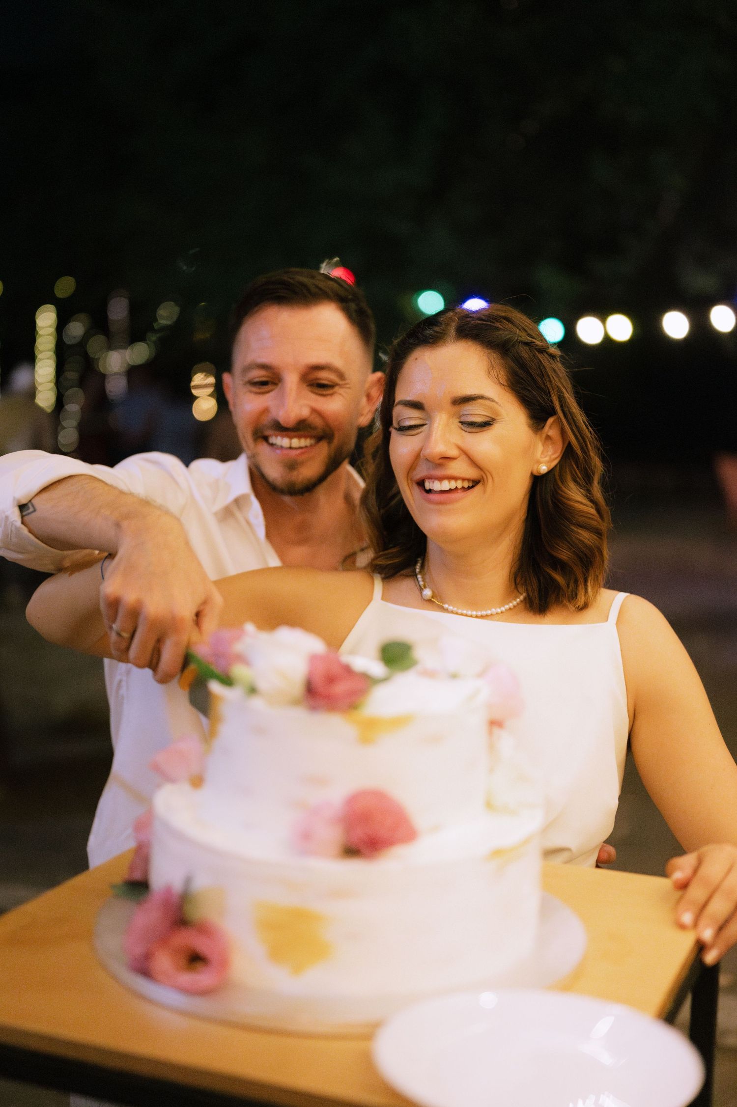 A couple shares a joyful moment while cutting into an elegant white wedding cake decorated with pink flowers at night.
