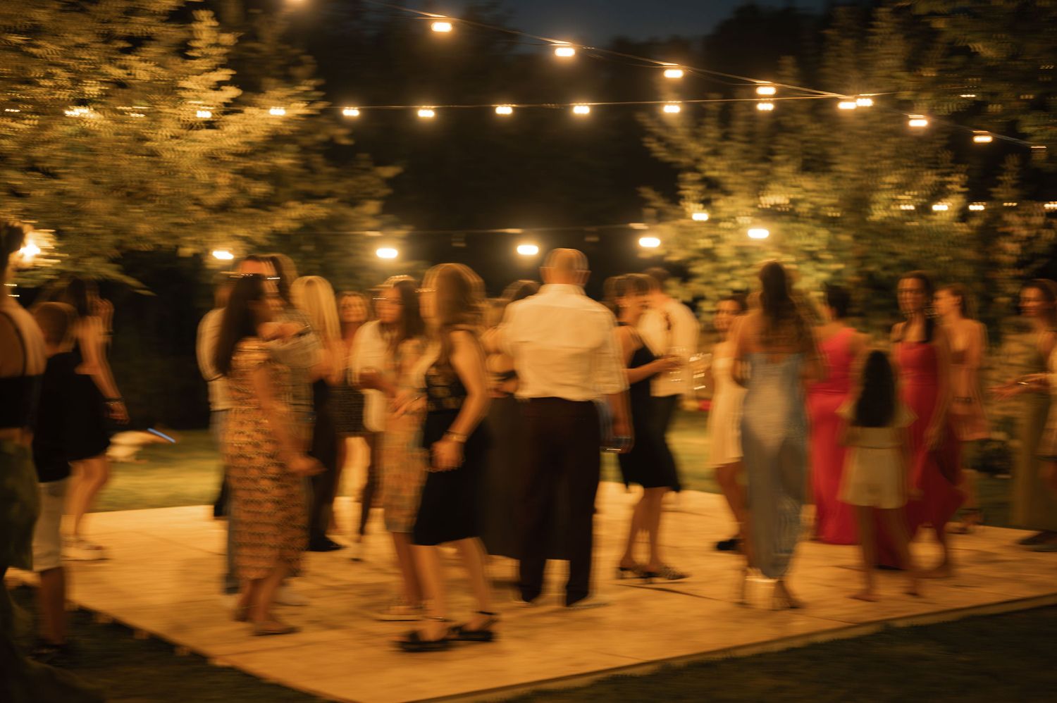 People dancing and socializing at an outdoor evening event with string lights illuminating the festive atmosphere.