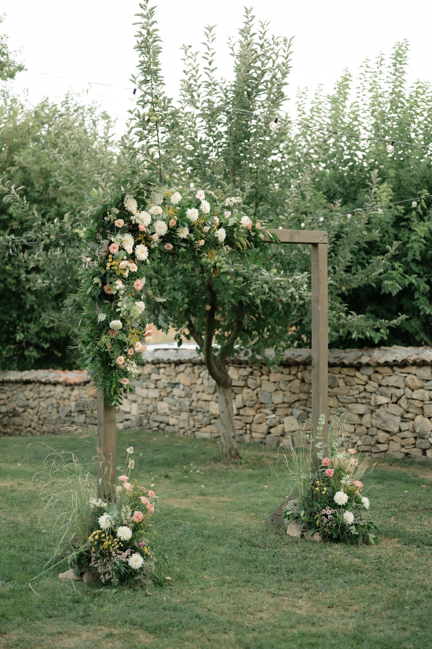 Floral arches with climbing roses and greenery line a stone wall in a garden setting.