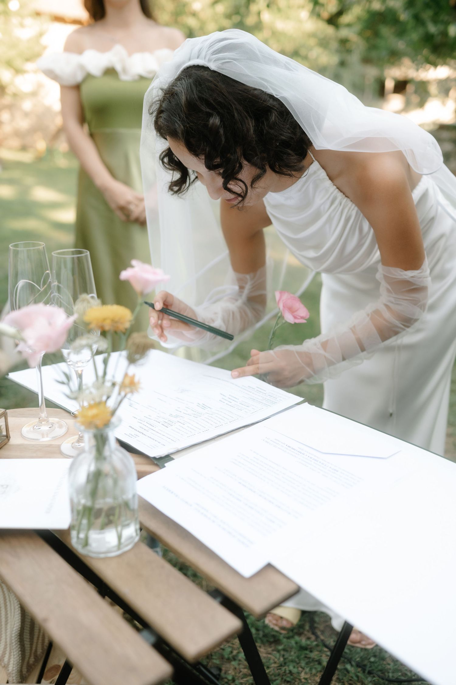 Someone signs a wedding guestbook at an outdoor reception with pink and yellow flowers in glass vases on the table.