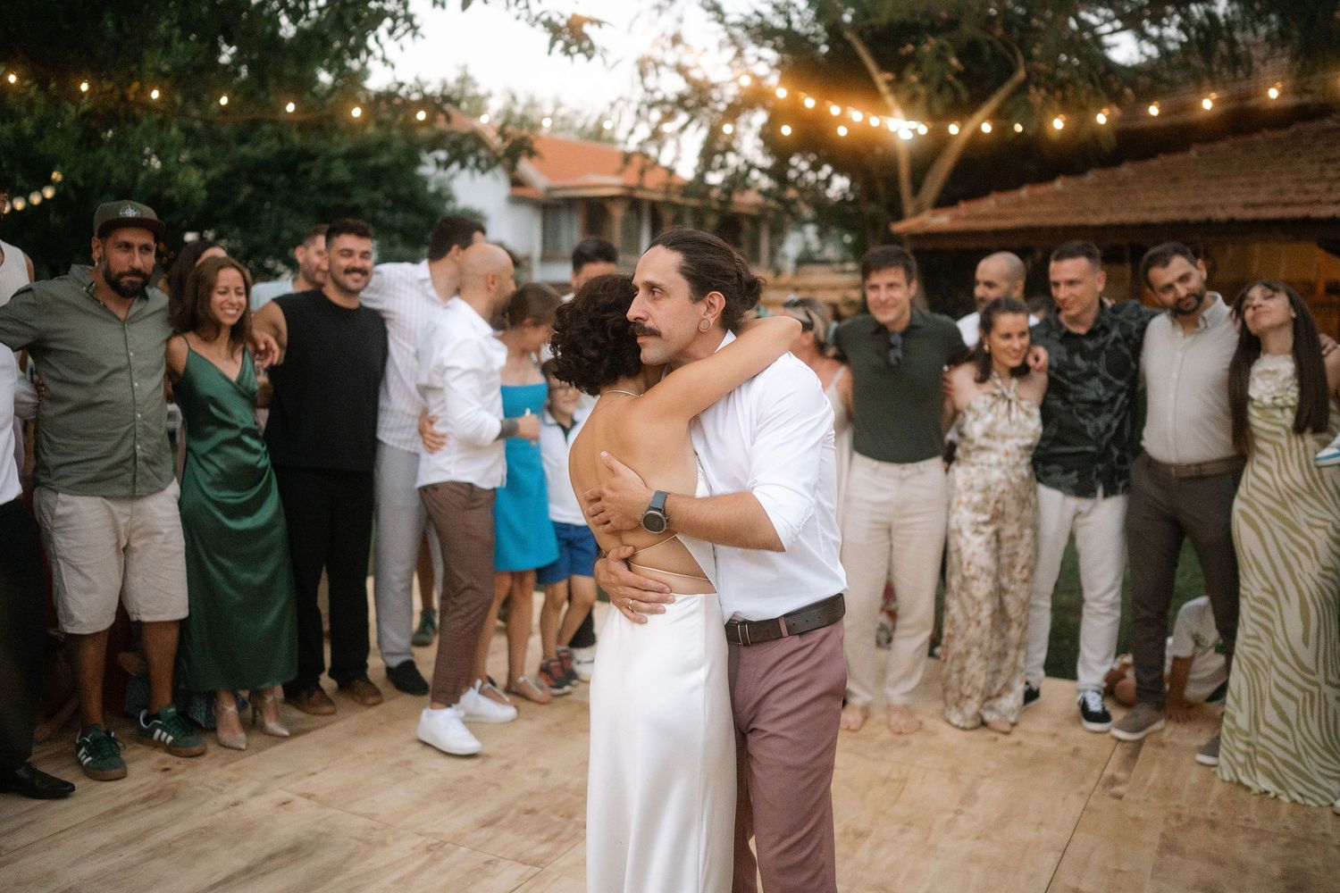 A group of wedding guests dance in a circle during an outdoor reception at sunset.