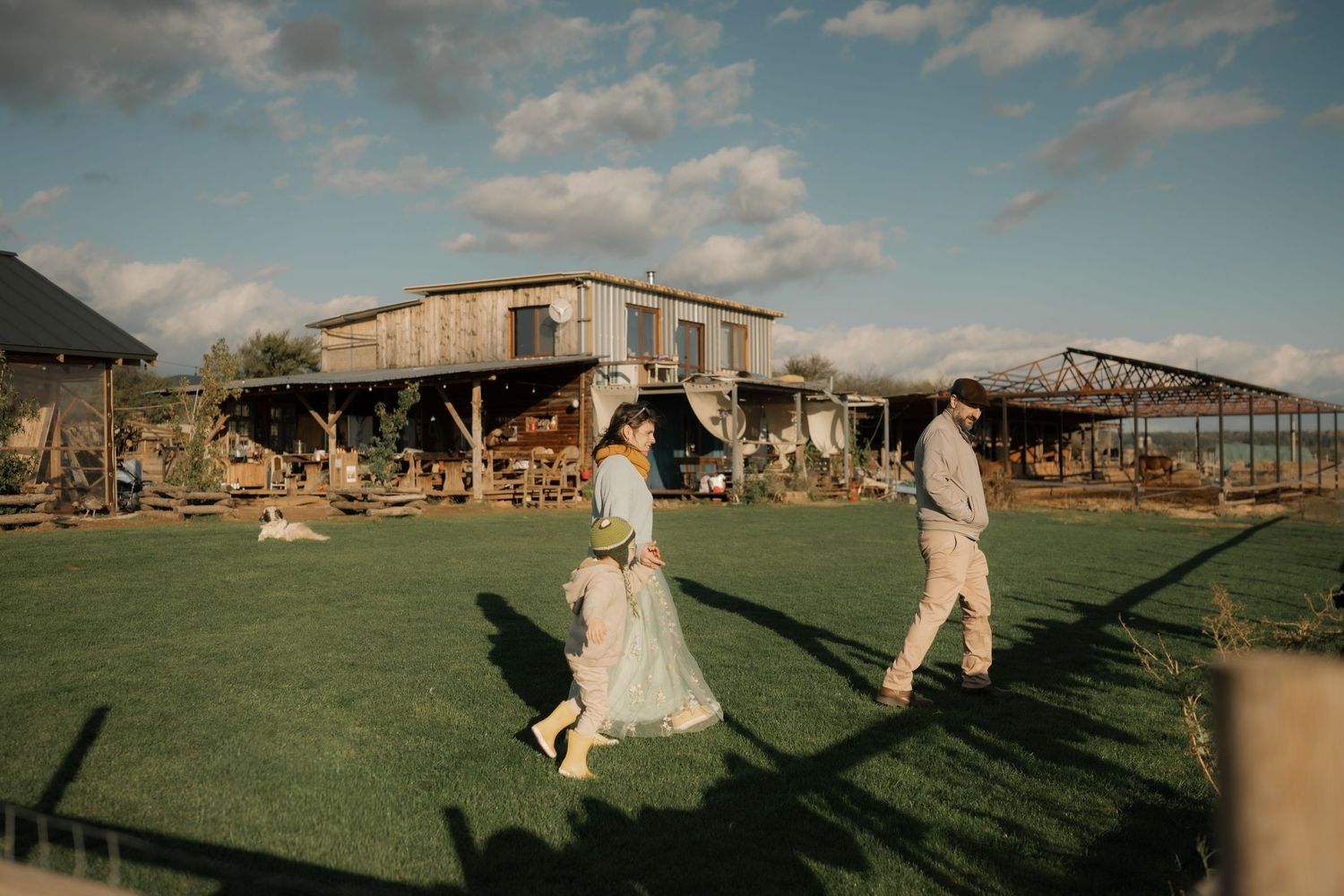 Bride and groom walk together casting long shadows on a grassy field during golden hour at a rustic outdoor venue.