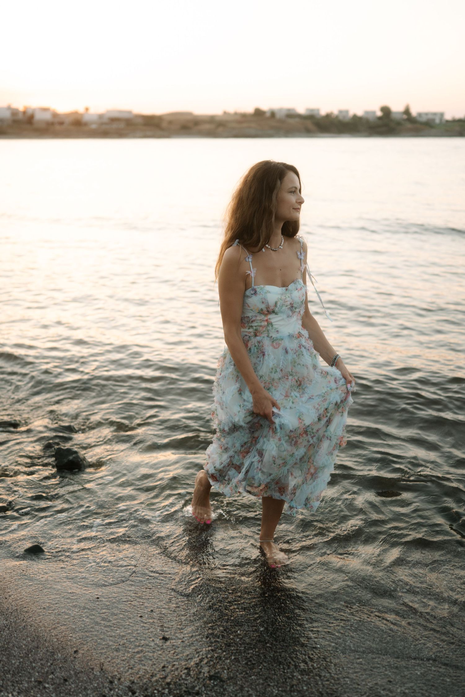 A figure in a floral dress walks along the shoreline at sunset creating ripples in the shallow water.