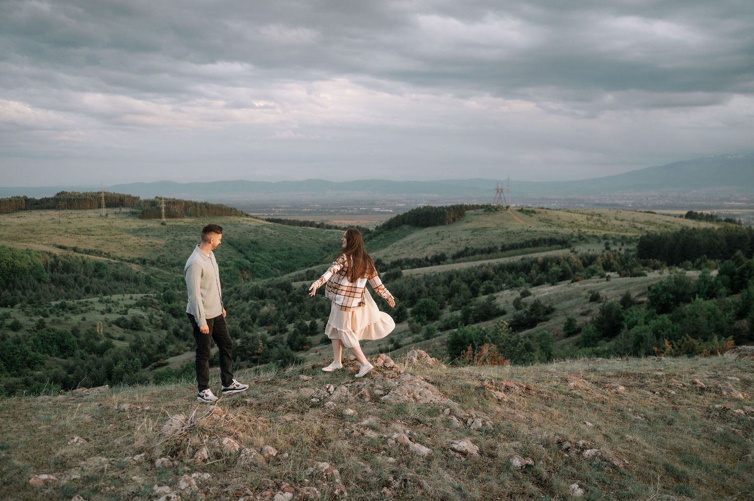 Two people walking on a hilltop overlooking a scenic valley landscape under moody cloudy skies.