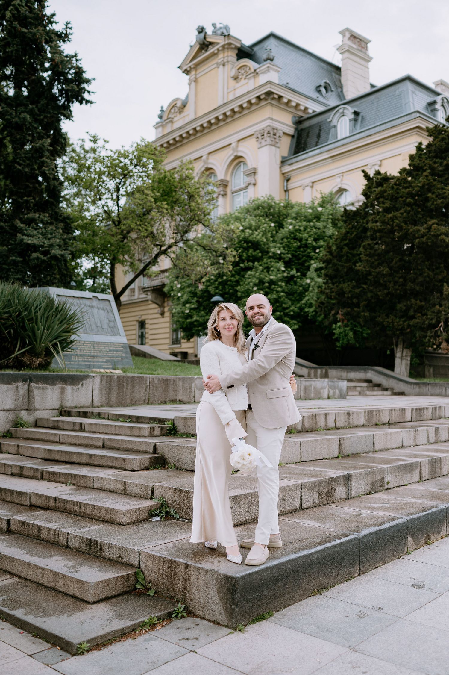 A couple in white outfits poses on stone steps with a grand historic building in the background.