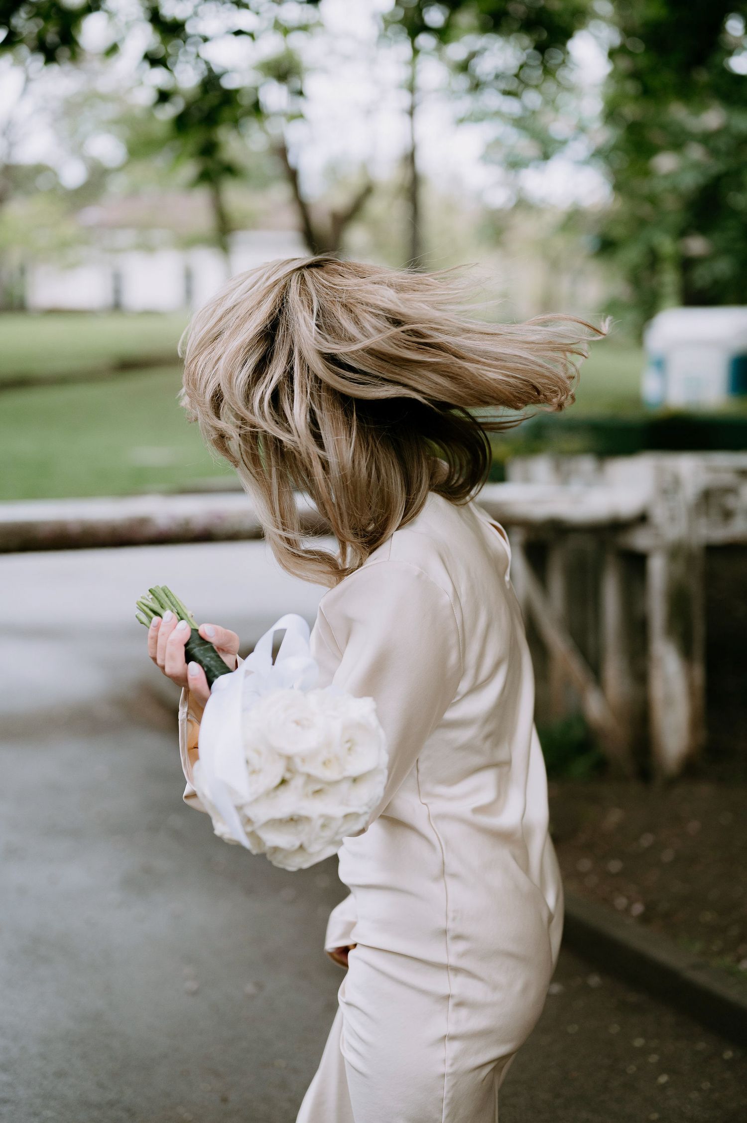 A person in a cream outfit with windswept hair holds a bouquet outdoors.