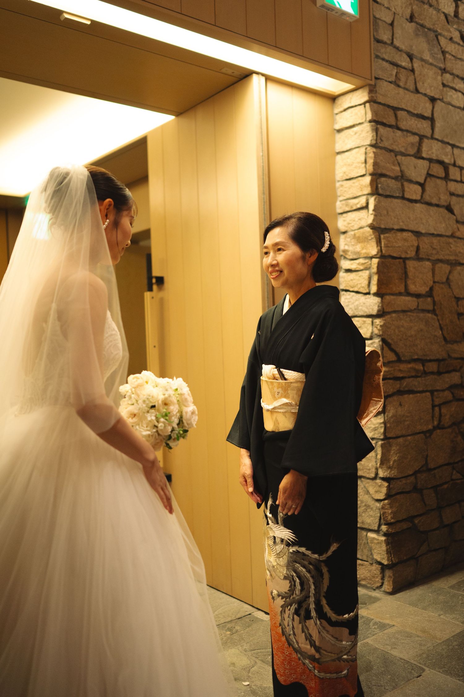 Bride in flowing white gown meets guests in an elegant hallway with stone accents.