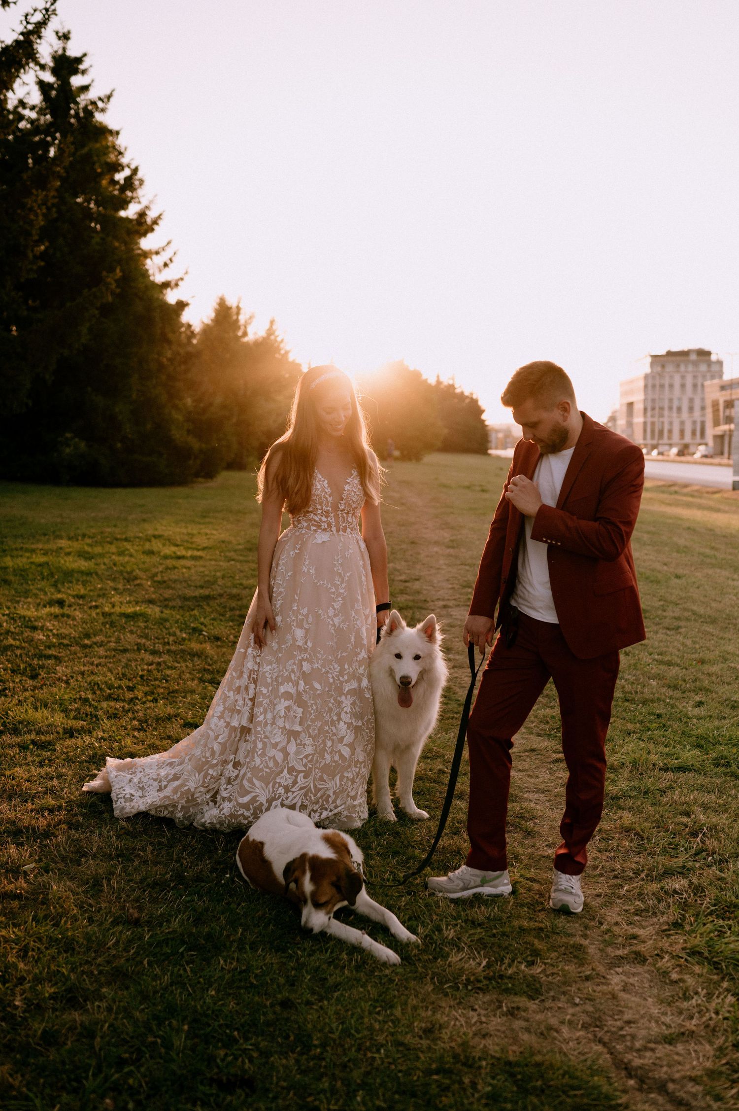 A couple poses with their white dog on a grassy hill during sunset with a building in the background.