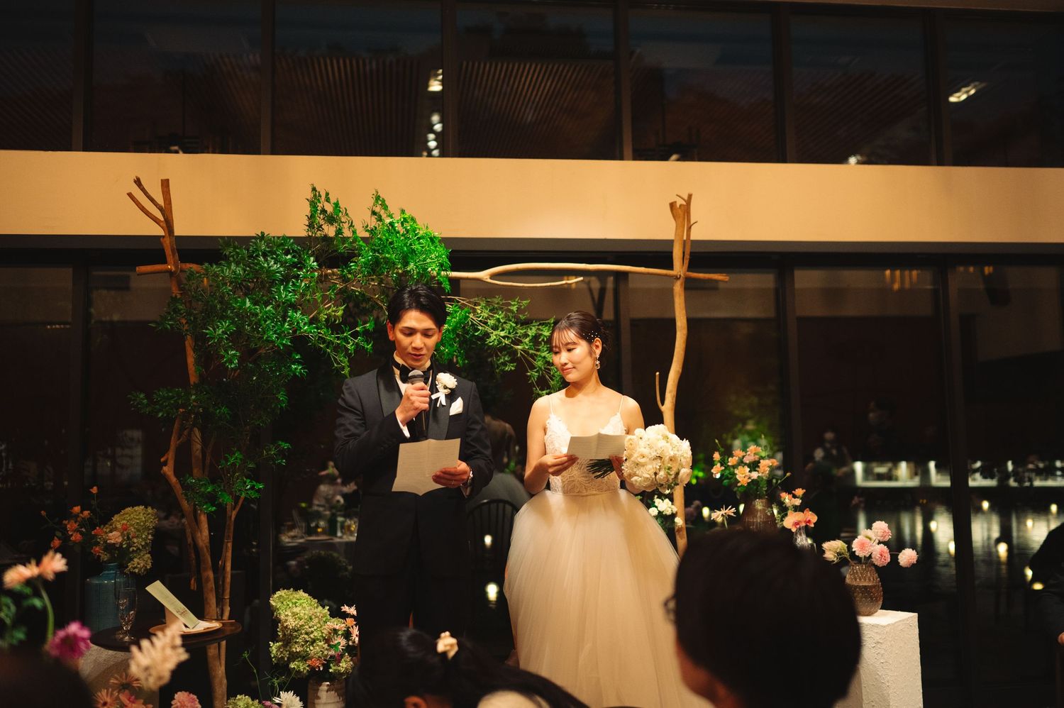 A couple celebrates during their wedding reception with raised hands under decorative lighting and greenery.