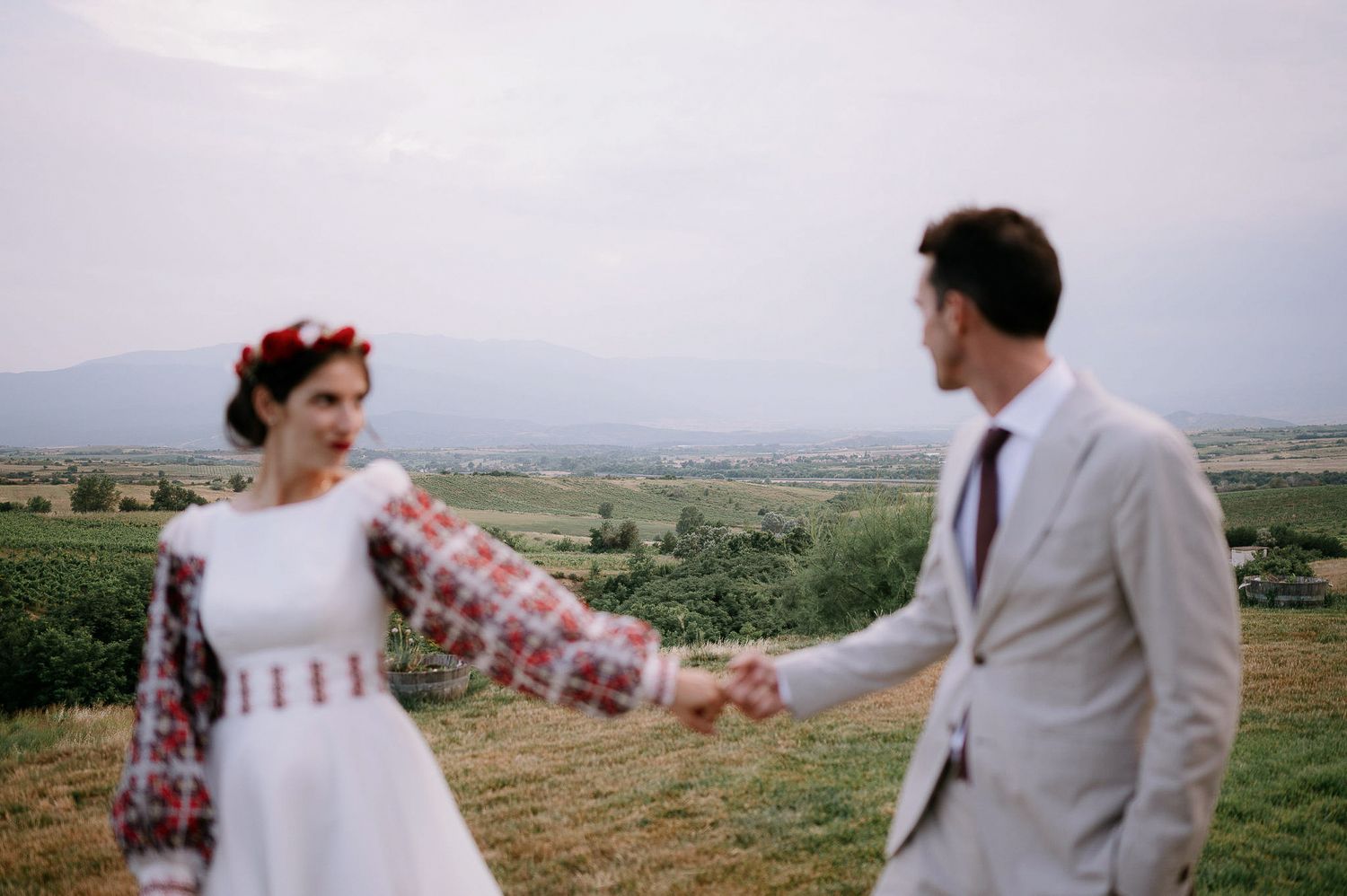 Wedding couple in folk-inspired dress and light suit stand together on a scenic overlook.