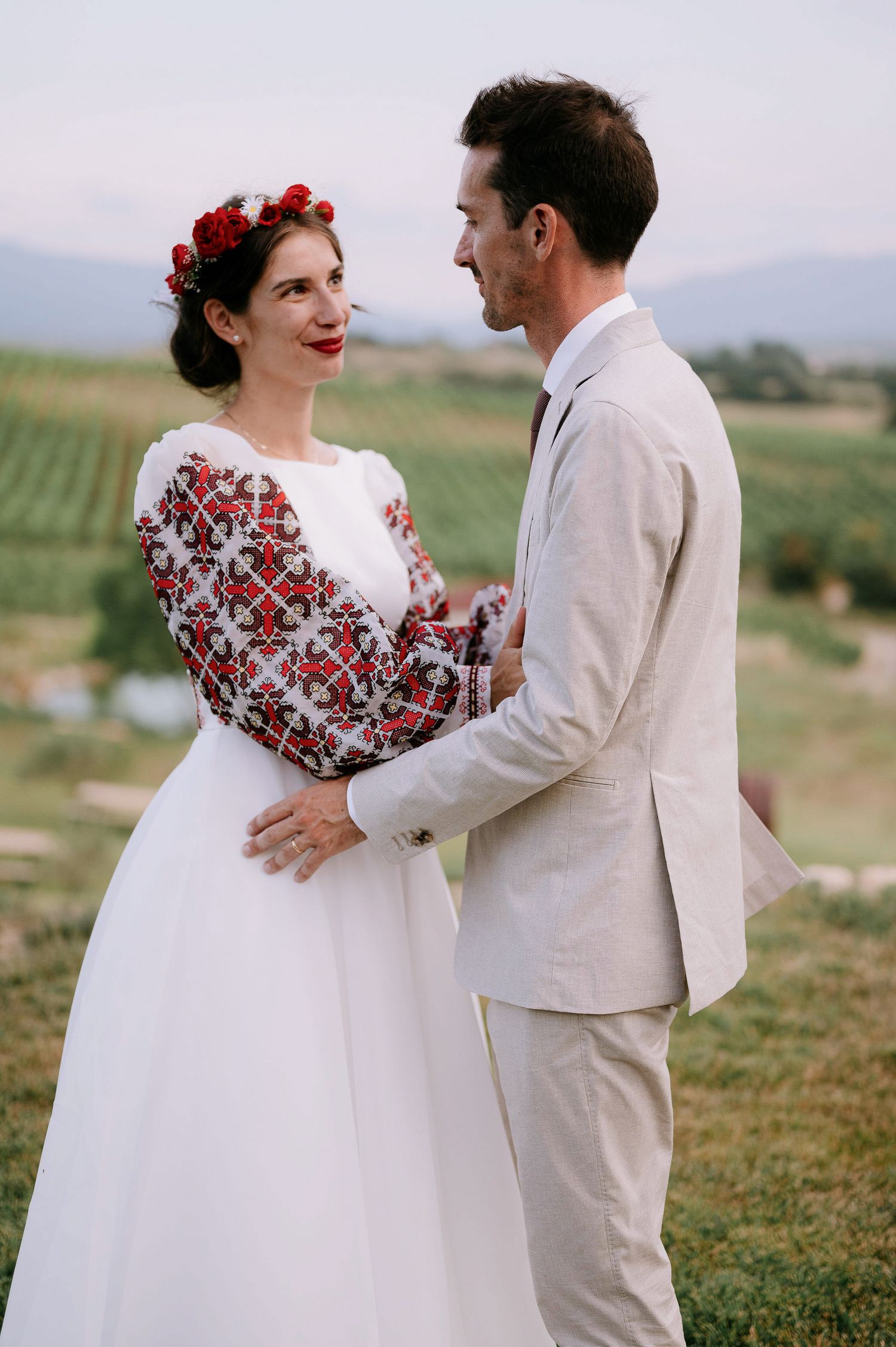 Wedding couple in traditional dress with unique red embroidered details share a joyful moment outdoors.