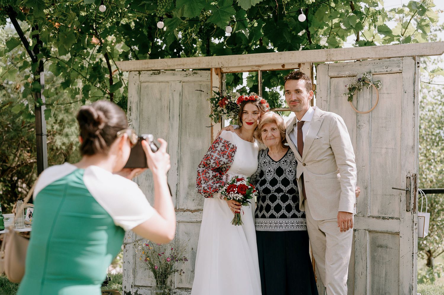 A photographer captures a wedding portrait in front of rustic white doors and greenery.