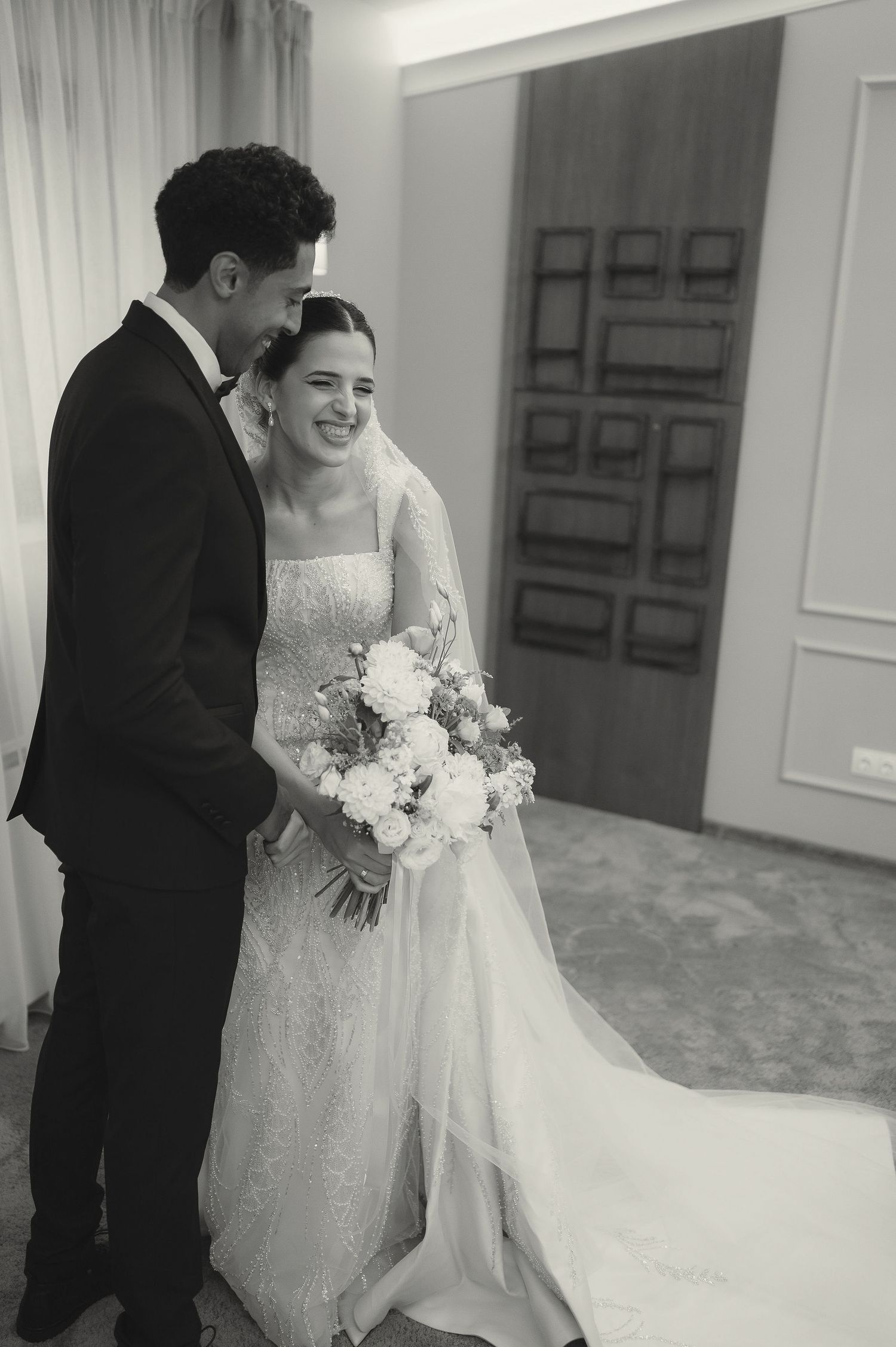 A couple in wedding attire shares an intimate moment in an elegant hallway with white walls and wooden doors.