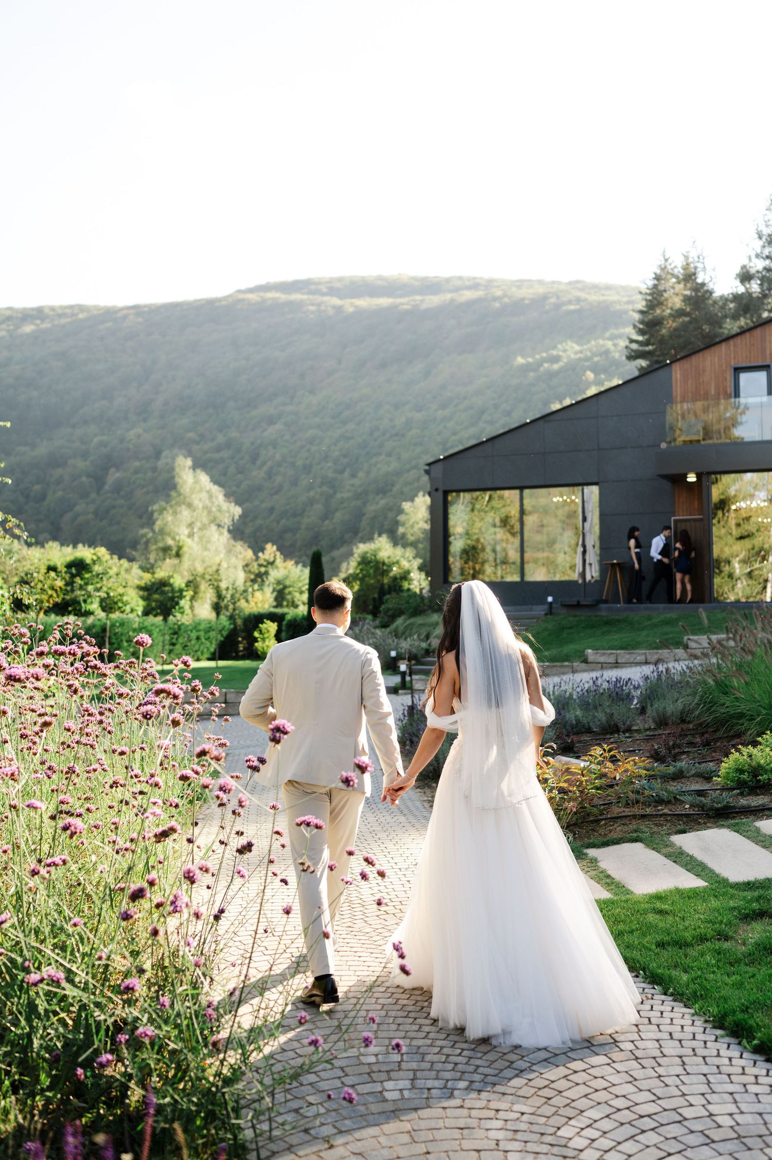 Wedding couple strolls along a mountain path with scenic valley views and blooming flowers.