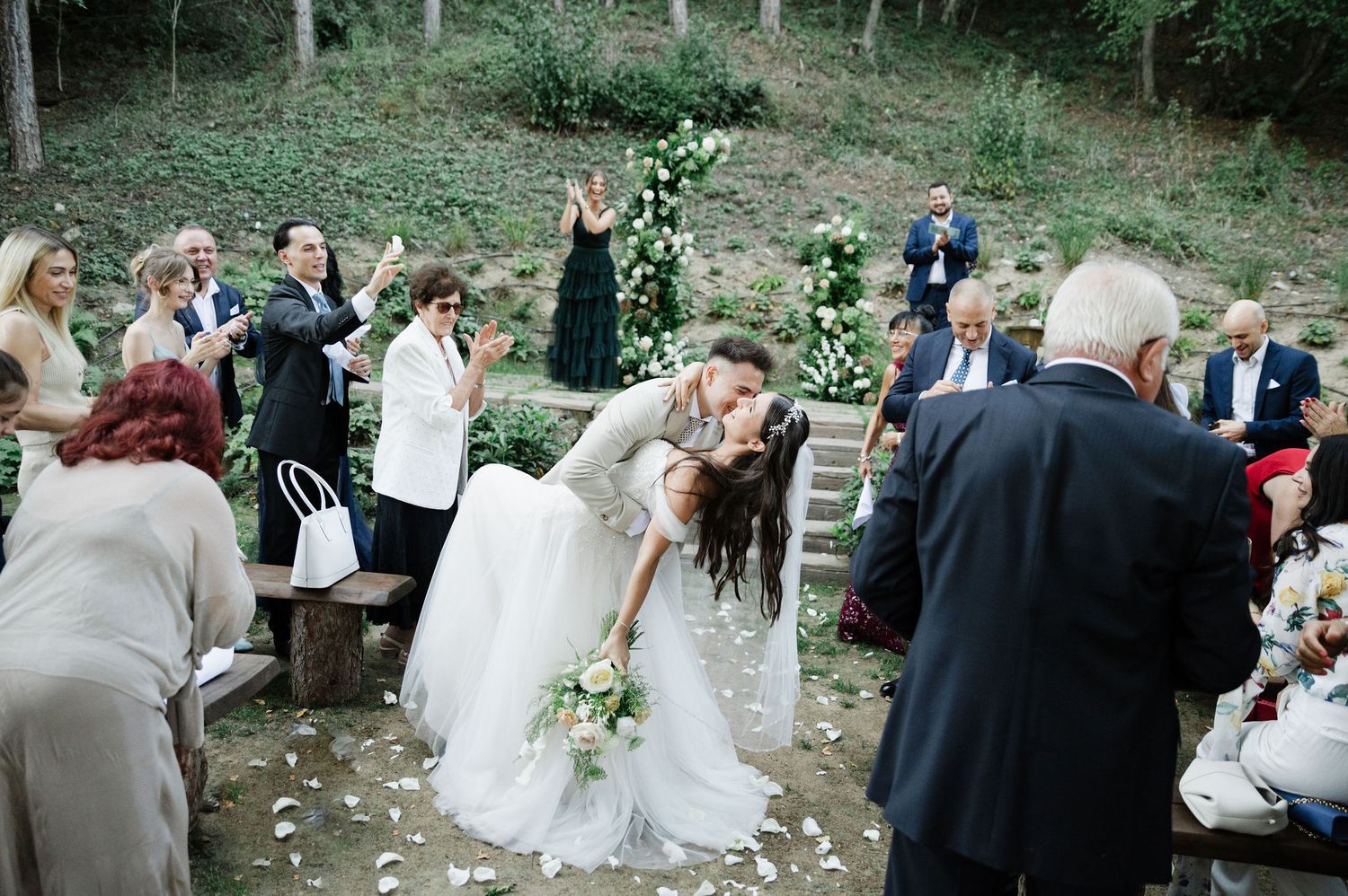 Outdoor wedding celebration with guests gathered in a wooded natural setting.