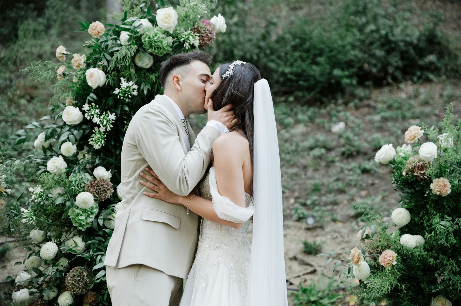 Romantic moment between newlyweds under rose-covered arch in forest.