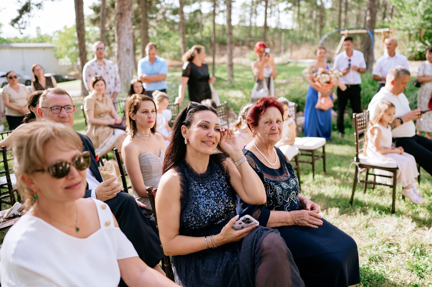 Wedding guests seated outdoors watch the ceremony among pine trees.