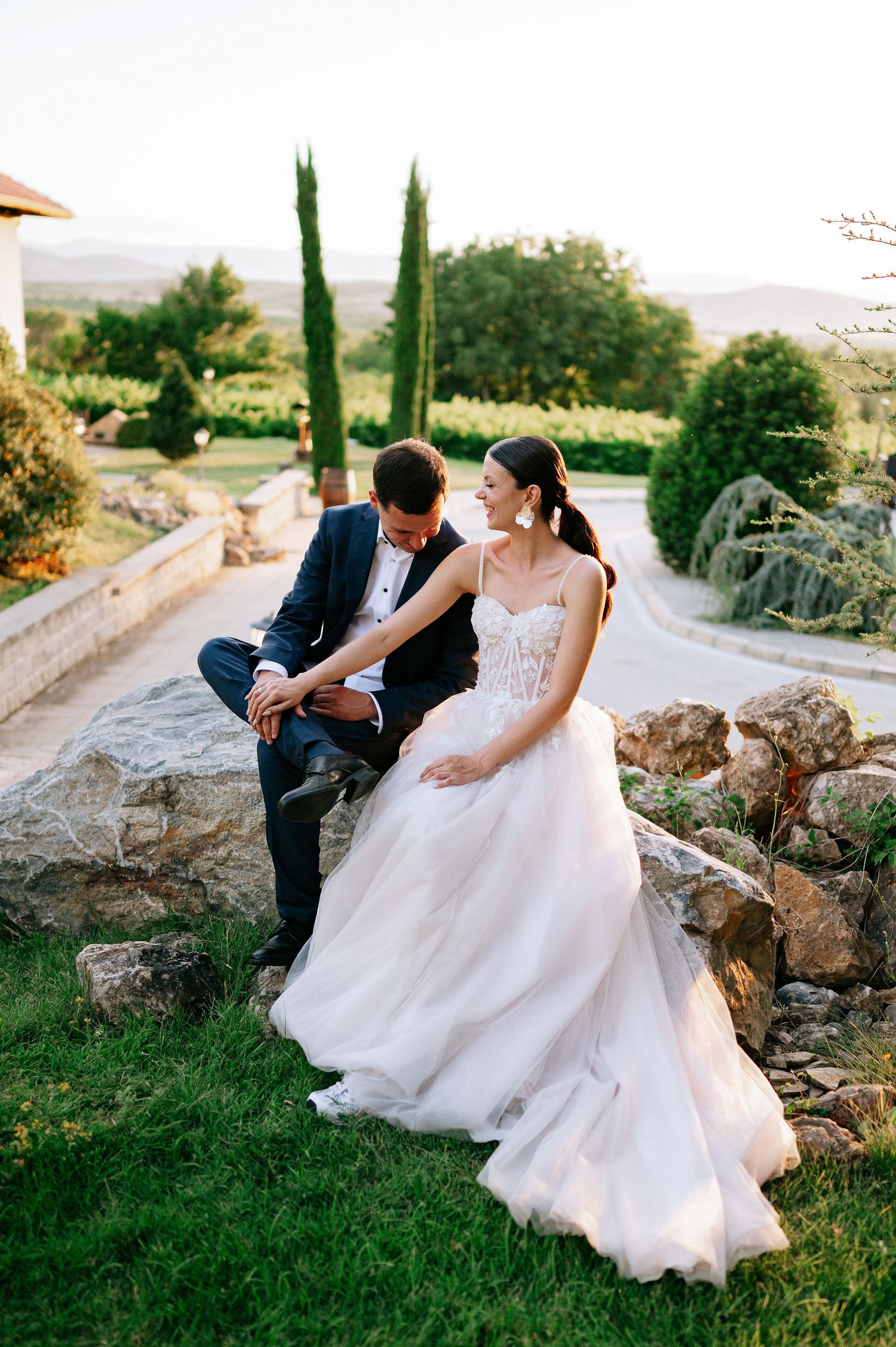 Wedding couple shares candid moments while seated on rustic rocks in an outdoor garden setting.
