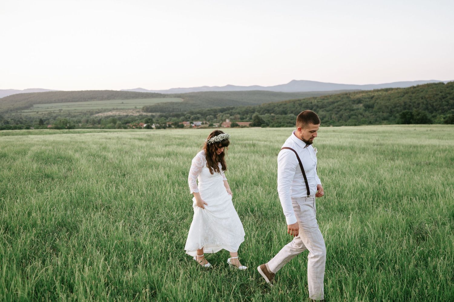 A romantic couple strolls through a lush green field against a mountainous landscape.