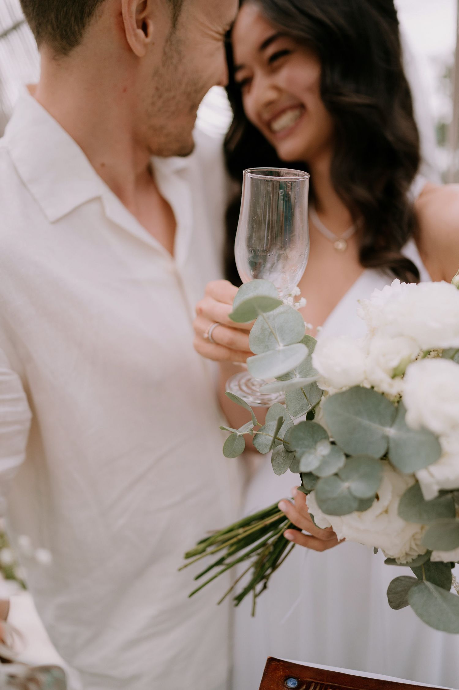Elegant wedding couple holding champagne glasses with white roses and eucalyptus bouquet.