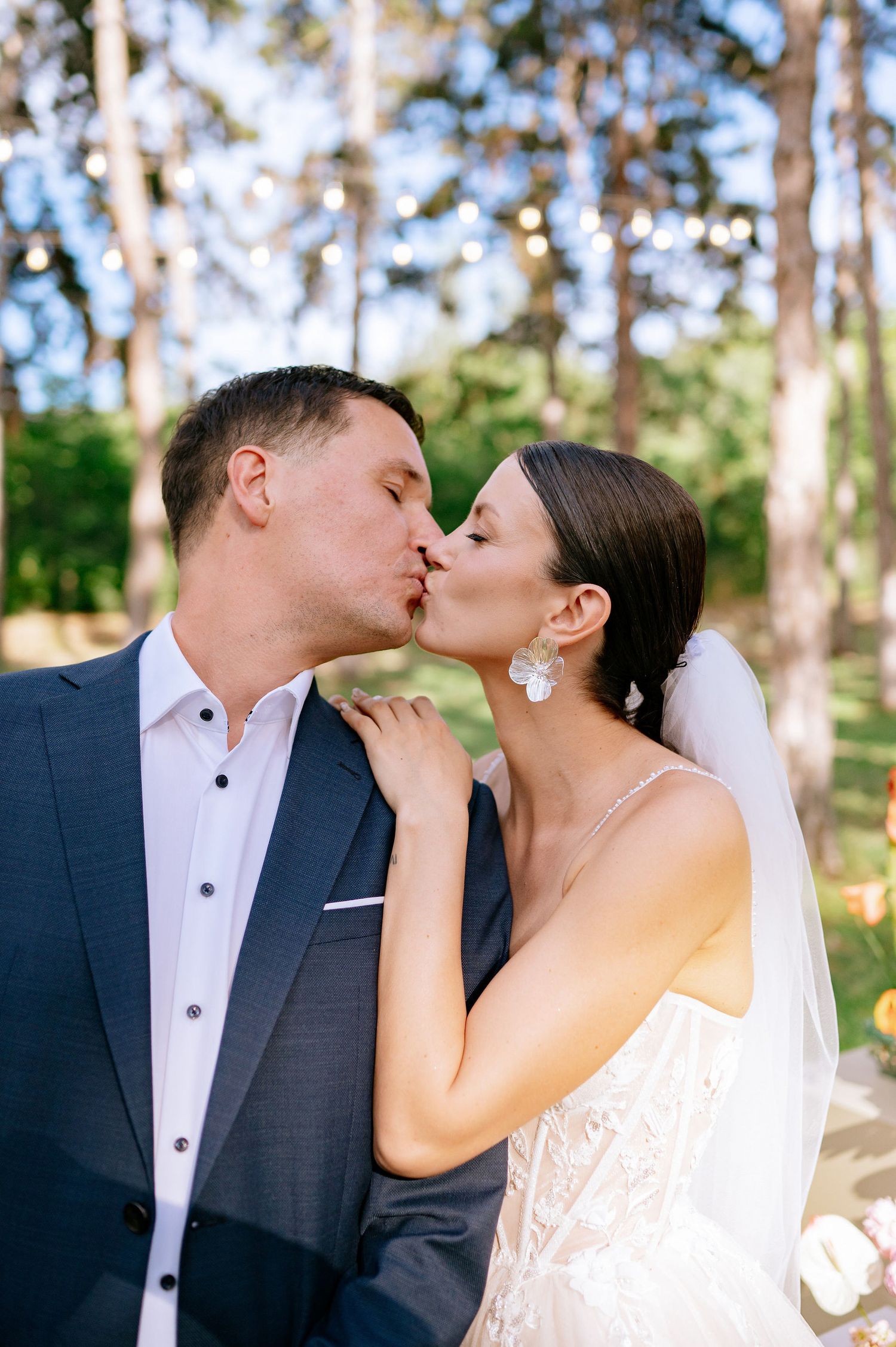 A tender wedding kiss photographed in a sunlit forest with floral decorations.