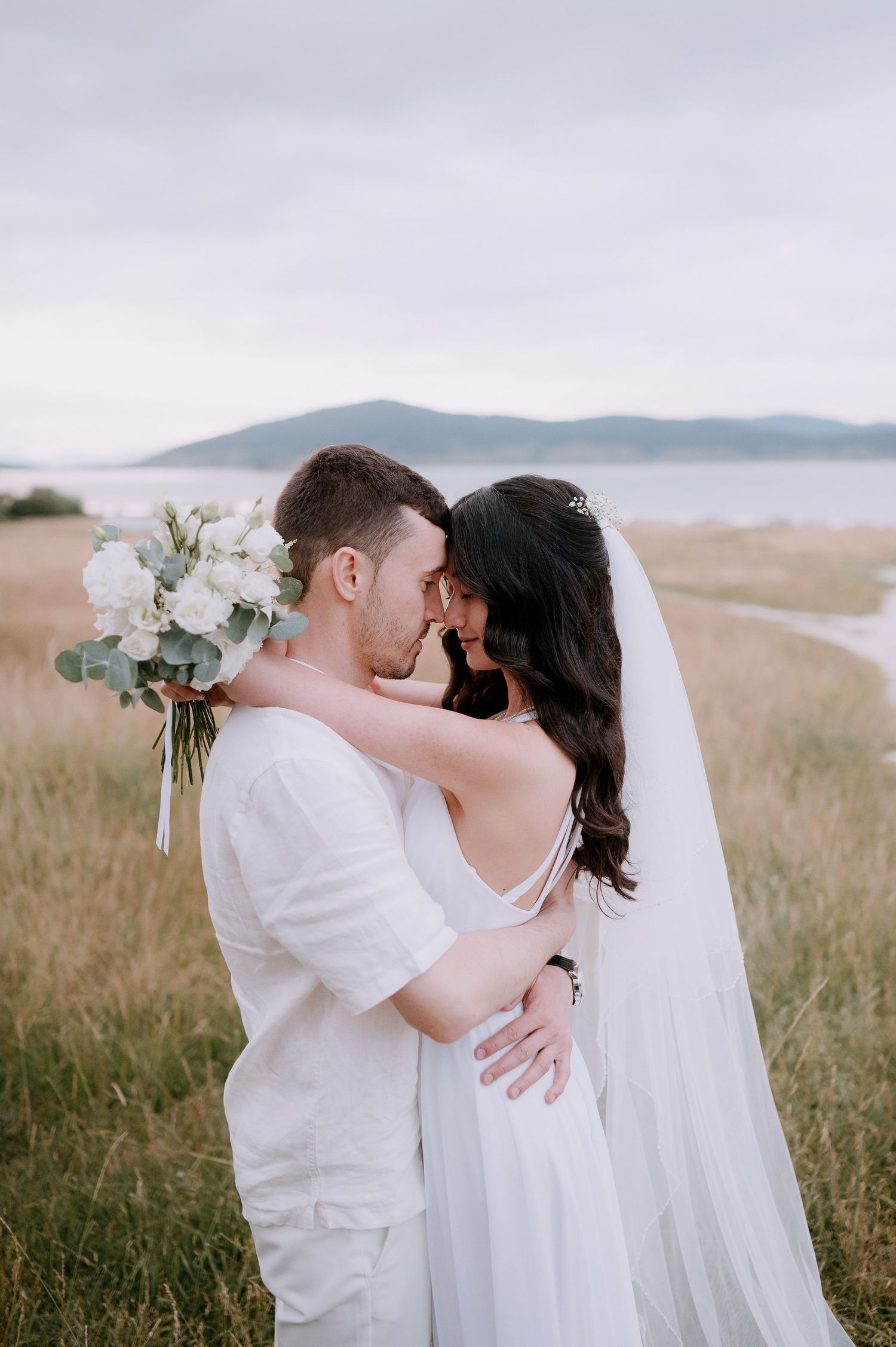 Intimate wedding moment captured in coastal field with flowing veil and white bouquet.
