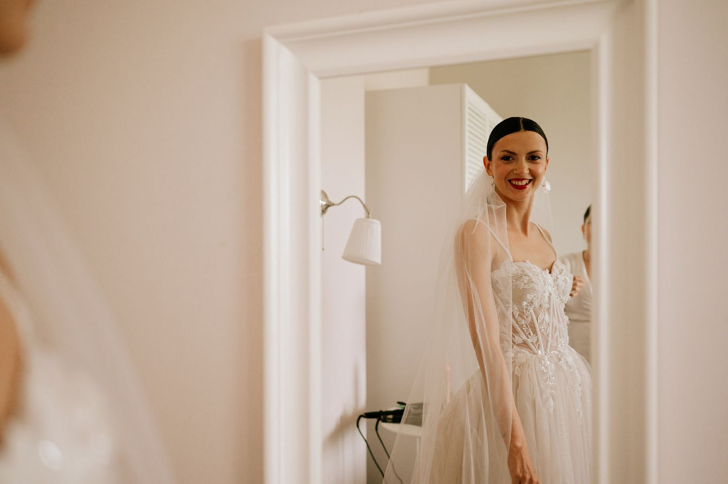 A bride in a white gown stands in an elegant room with natural lighting.