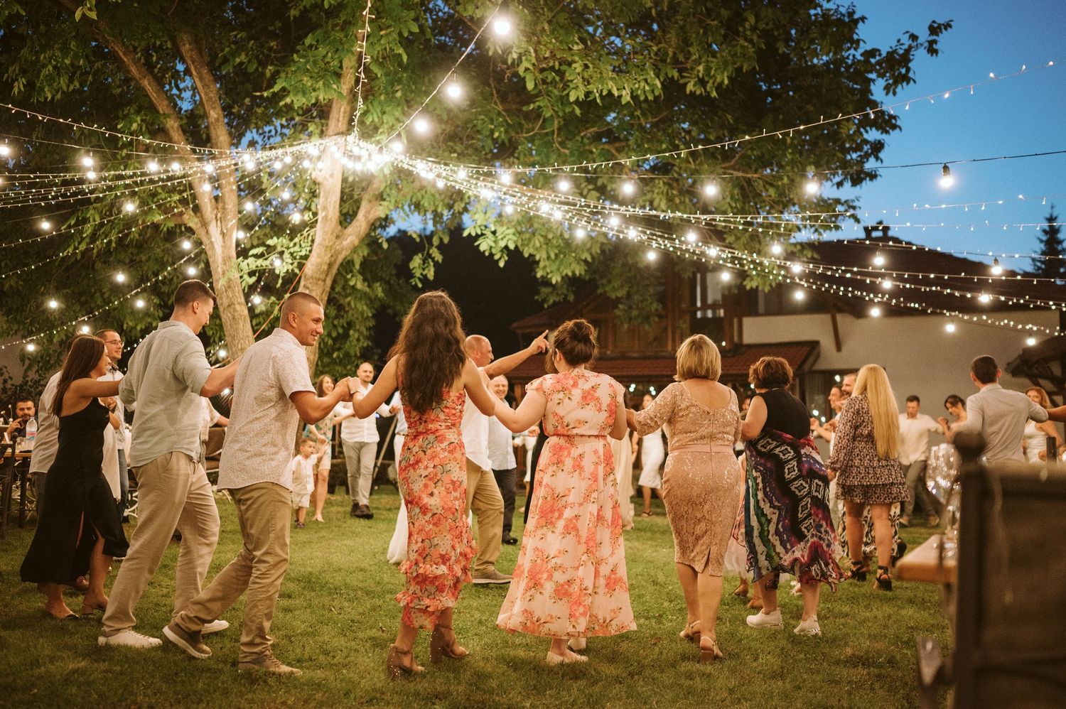 Wedding guests dancing in a garden setting illuminated by overhead string lights at dusk.