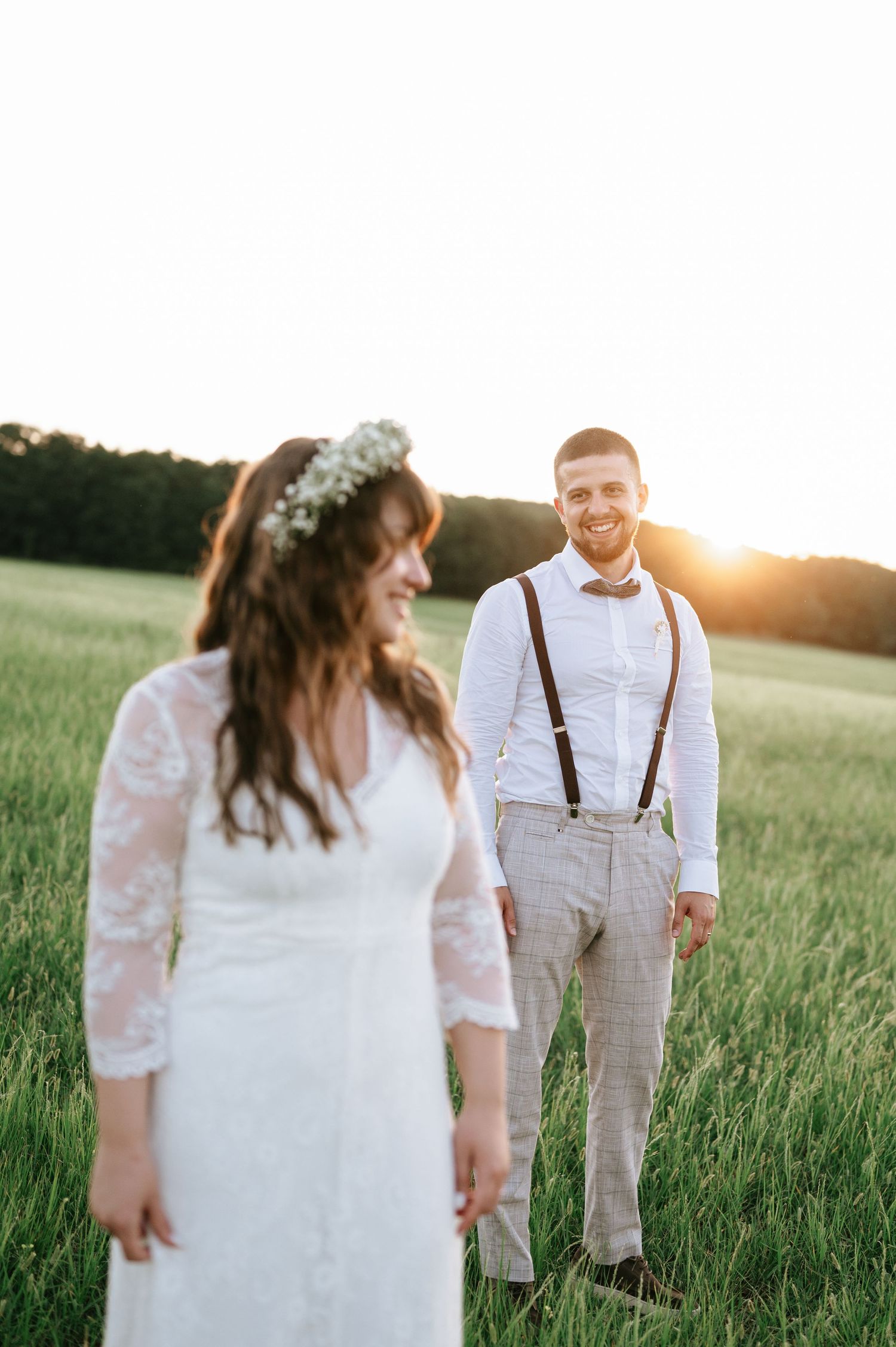 Sunset wedding portraits in a grassy field with bride in lace gown and groom in suspenders.