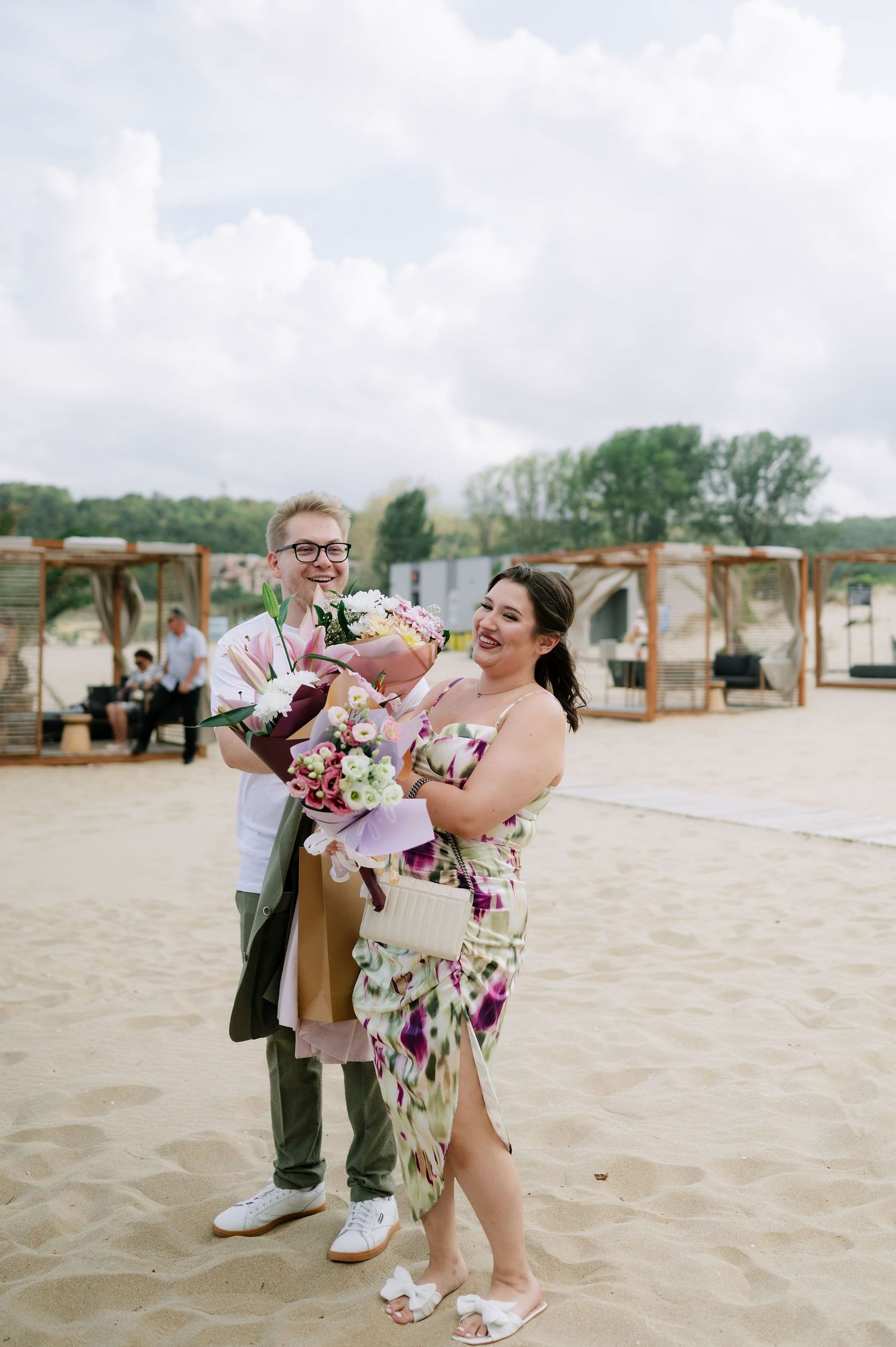 Couples dance together on a sandy beach with wooden structures in the background.